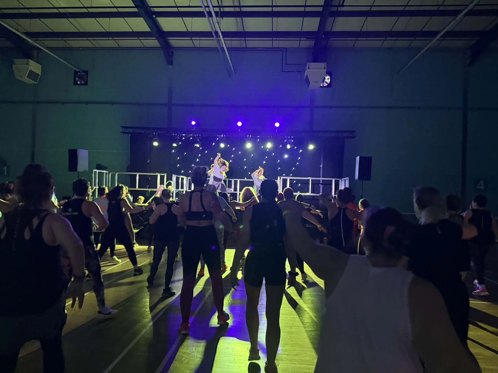 People dancing in a gymnasium with colorful stage lights, a stage with performers, and a high ceiling.