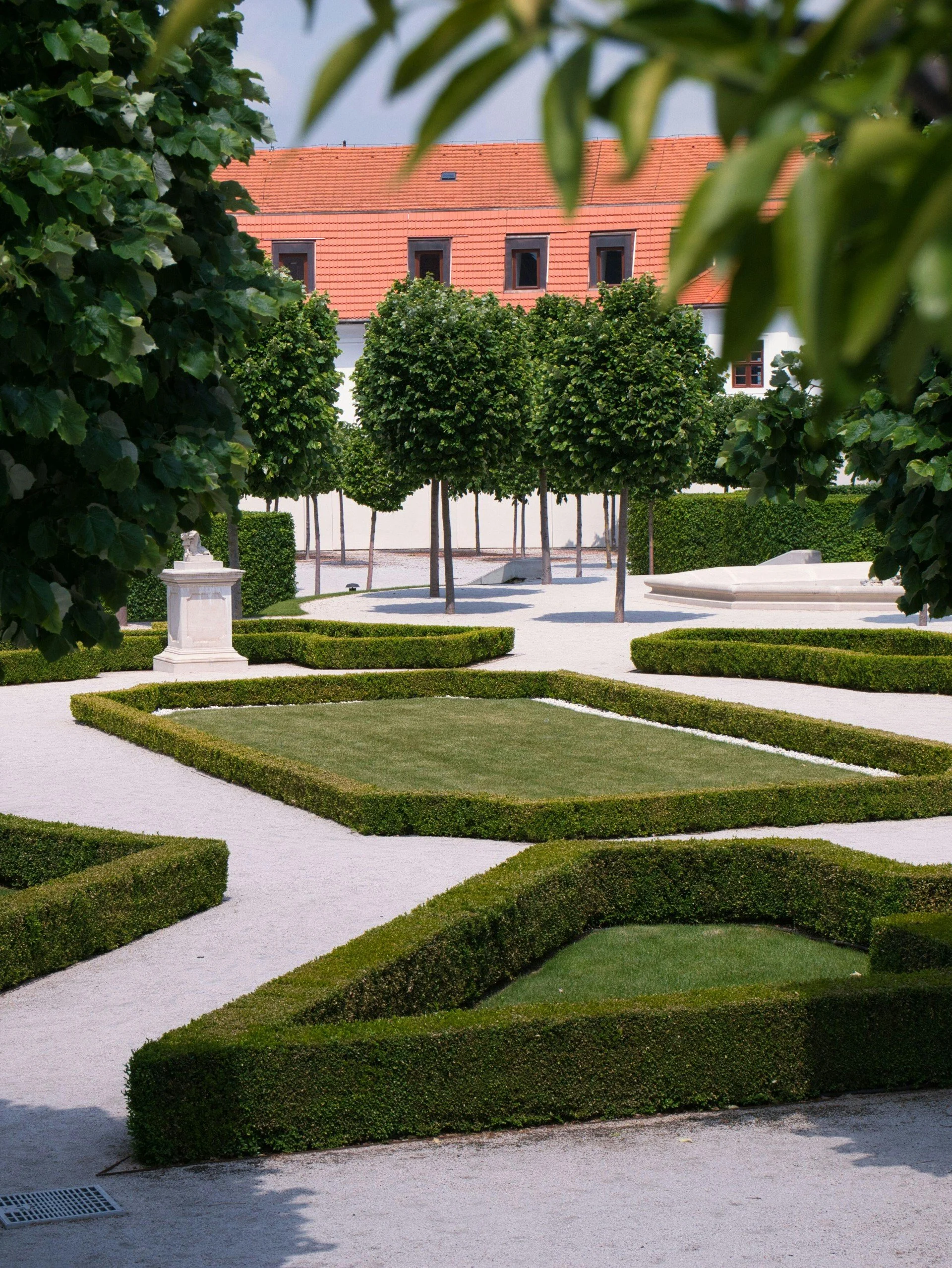 are & are Landscape Design Studio - Garden design with a asymmetrical parterre garden hedges, trees, gravel walkways, and classical-style sculptures, with a building featuring a red-tiled roof in the background.