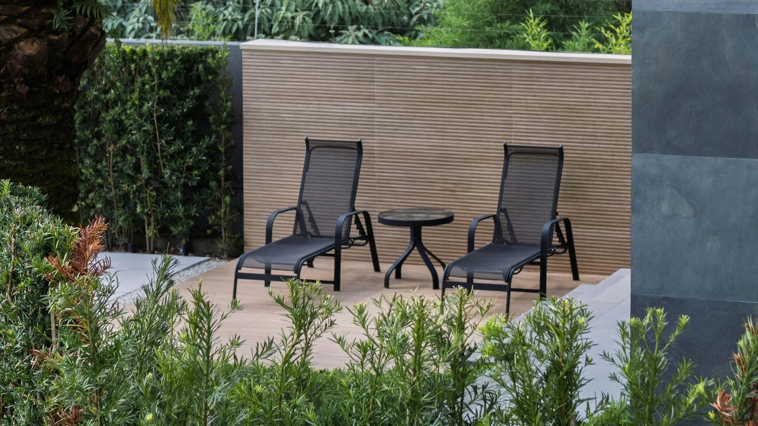 are & are Landscape Design Studio - Garden design with wo black outdoor chairs and a small round table on a wooden patio, surrounded by green shrubs and plants, with a textured beige wall behind and a dark gray wall on the side.