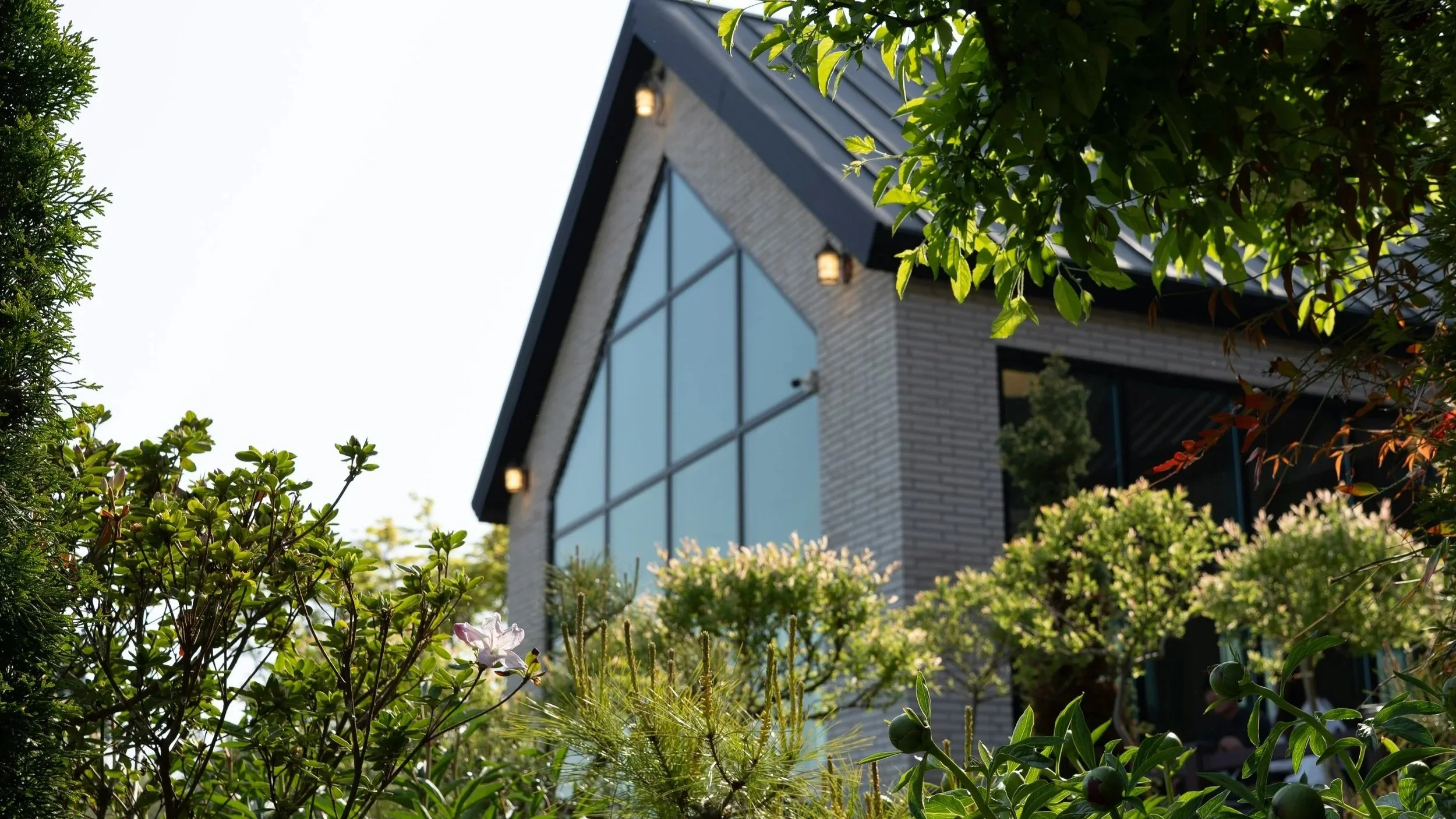 are & are Landscape Design Studio - Garden design with a modern house with a sloped roof, large glass windows, and external lights, surrounded by lush green trees and plants for landscaping.