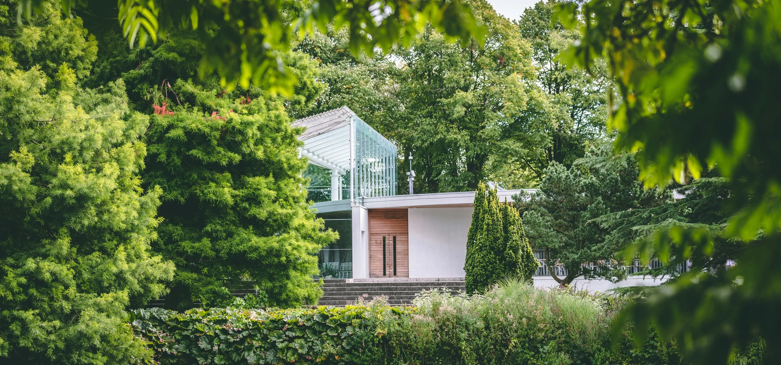 A modern house with a glass-walled balcony surrounded by lush green trees and plants in a garden setting.