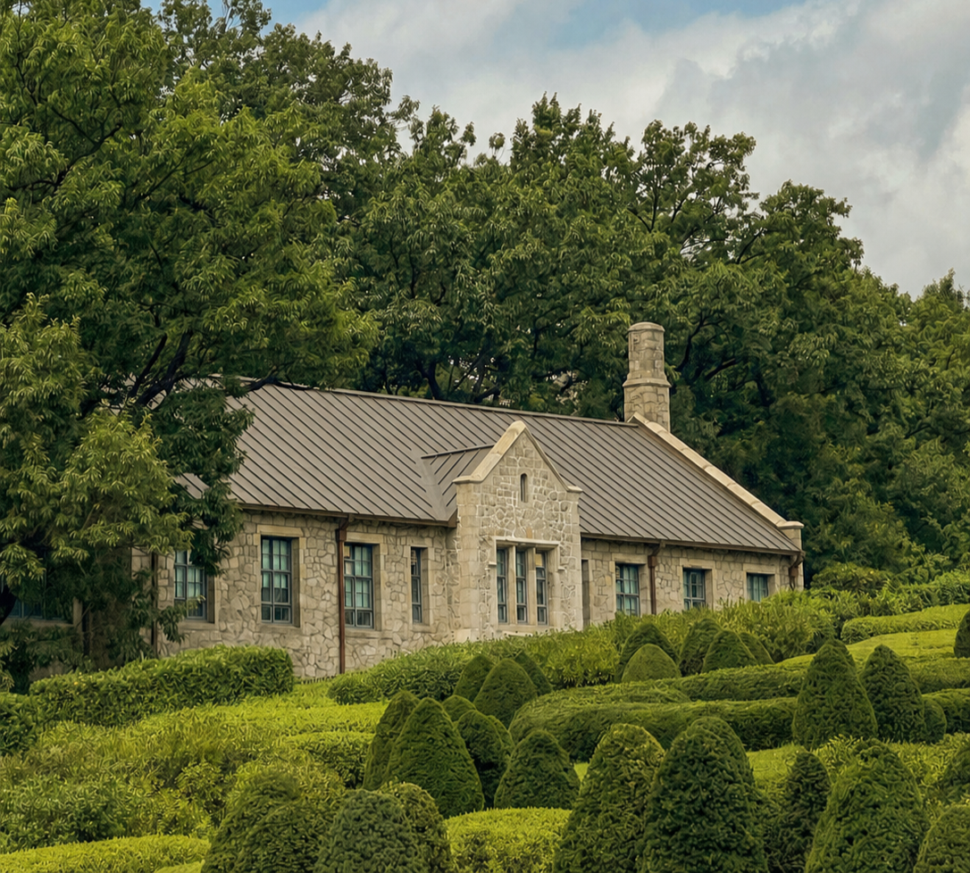 A formal landscaped garden with topiary hedges, mature trees and a stone house in the background.