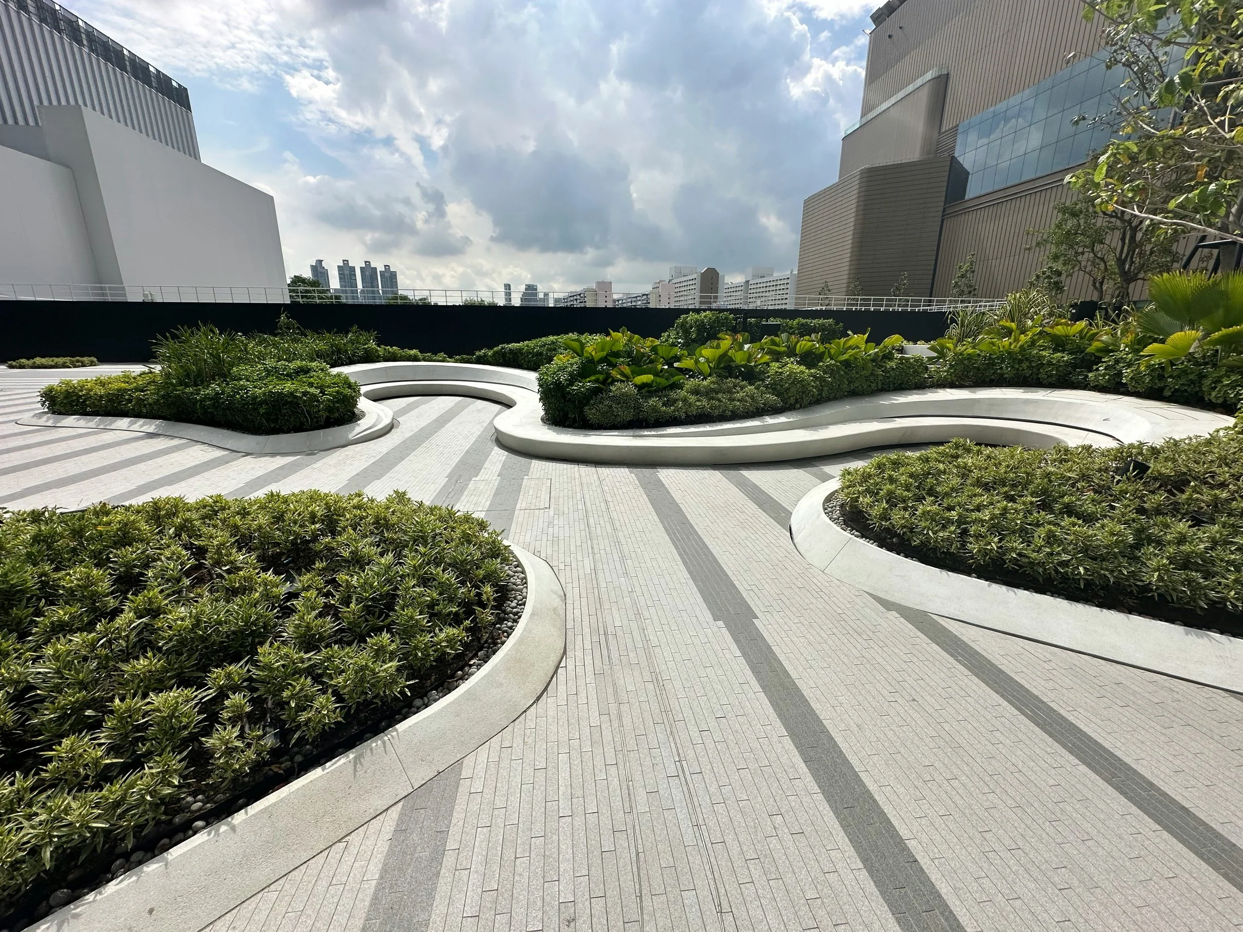 are & are Landscape Design Studio - Garden design - Urban rooftop garden with curved flower beds filled with green plants, surrounded by a tiled walkway, modern buildings, and a cloudy sky in the background.