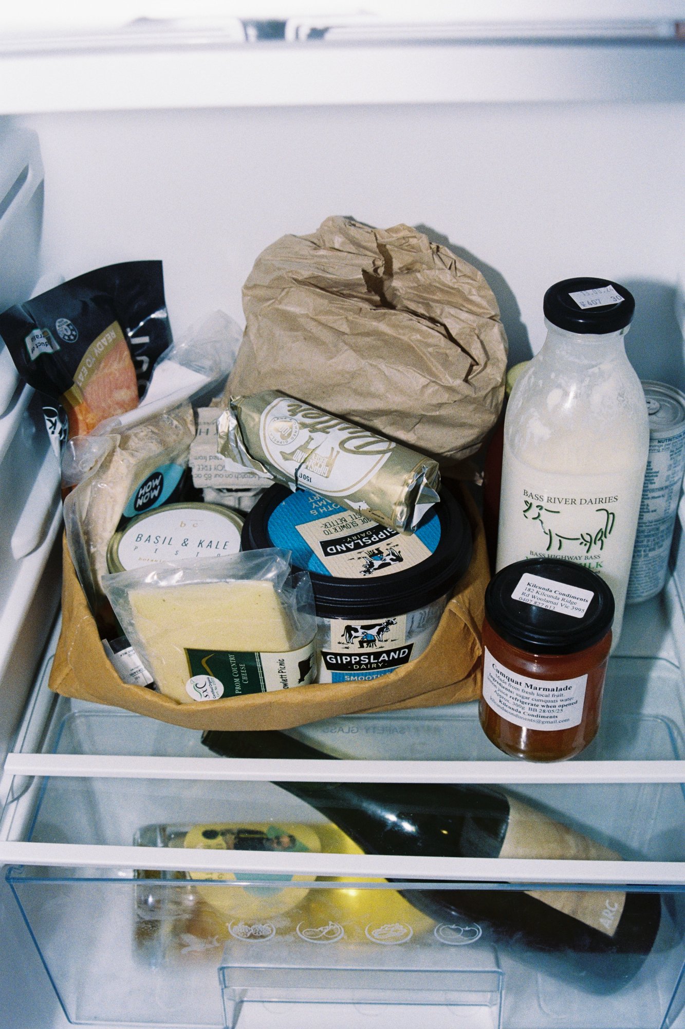 Inside of a refrigerator drawer with various food items, including a jar of yoghurt, a bag of snacks, a paper-wrapped item, a bottled drink, and other food containers.