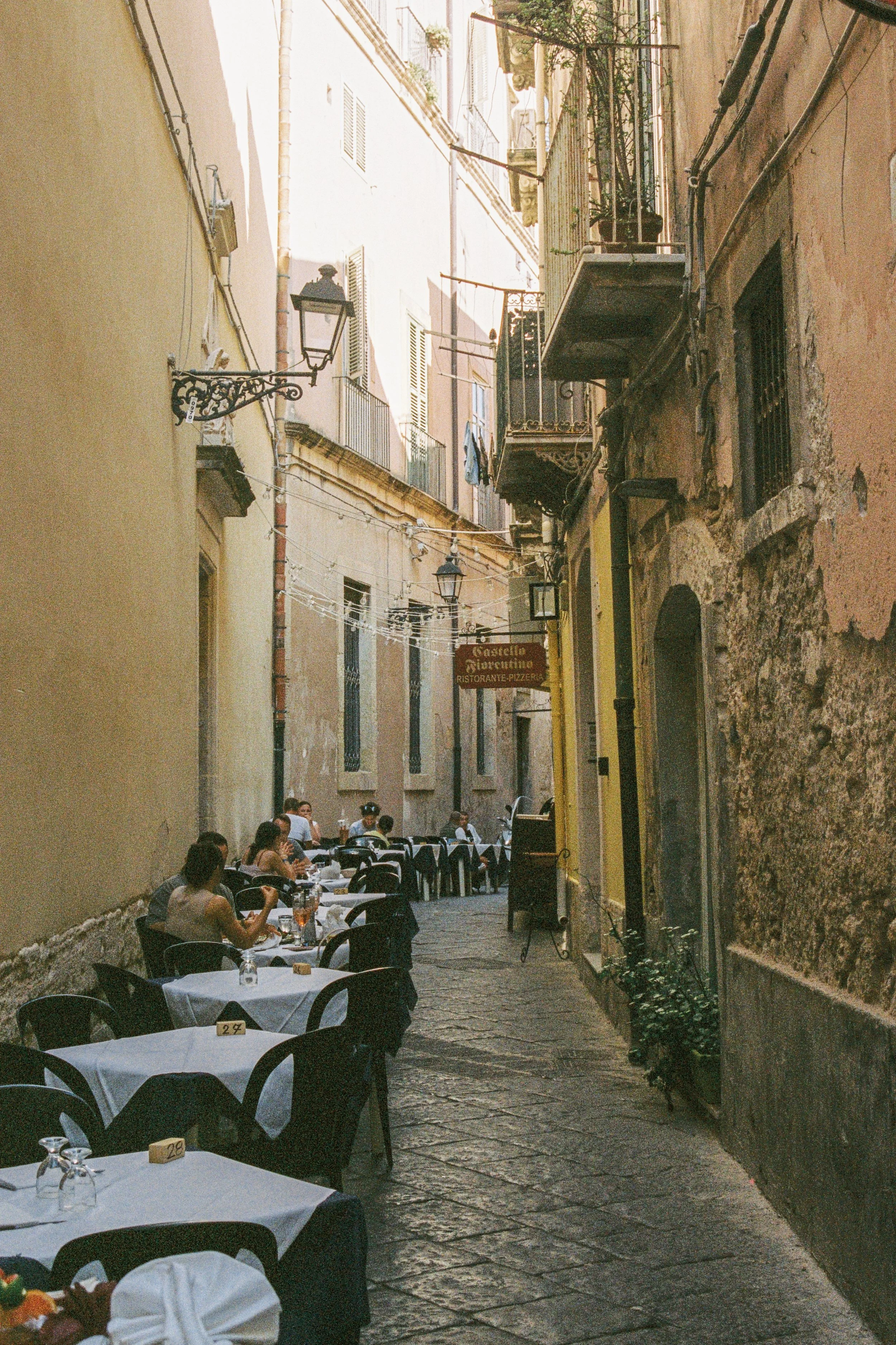 Outdoor dining area with tables and chairs along a narrow European street, with people sitting and enjoying food. Buildings with balconies and hanging plants line the street, and string lights are overhead.