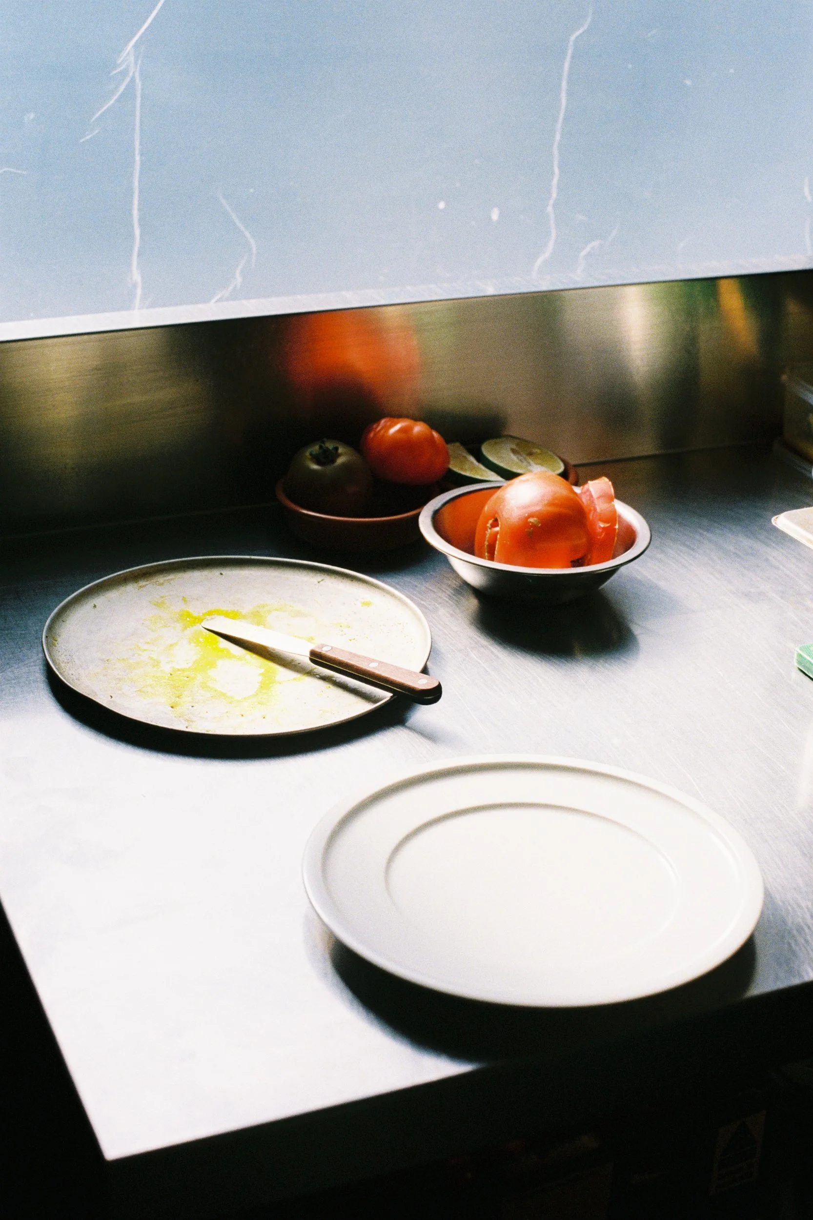A kitchen counter with various vegetables and plates, including tomatoes and lime slices, with a marble backsplash.