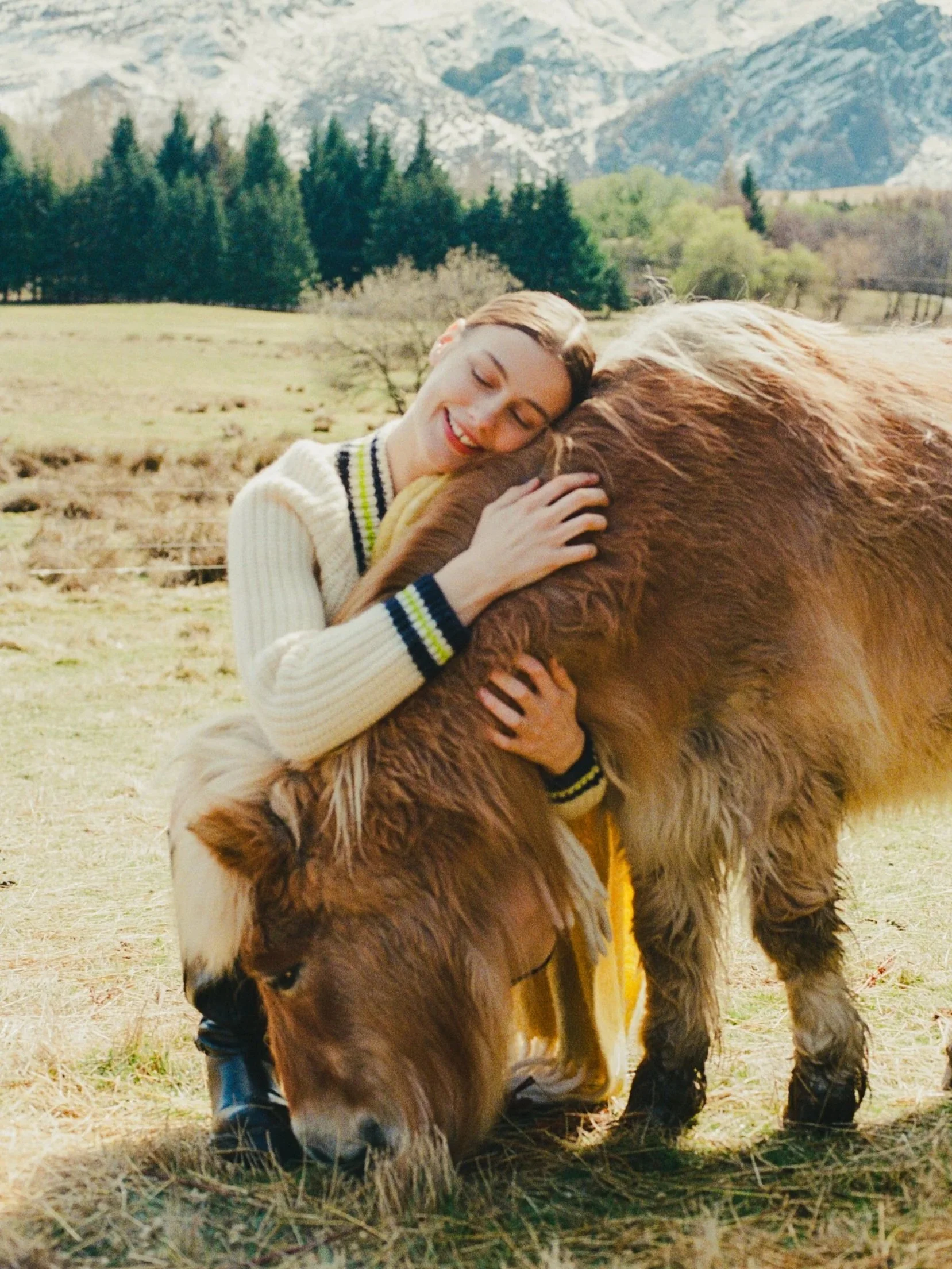Young woman hugging a large brown and white cow in a grassy field with mountains and trees in the background.