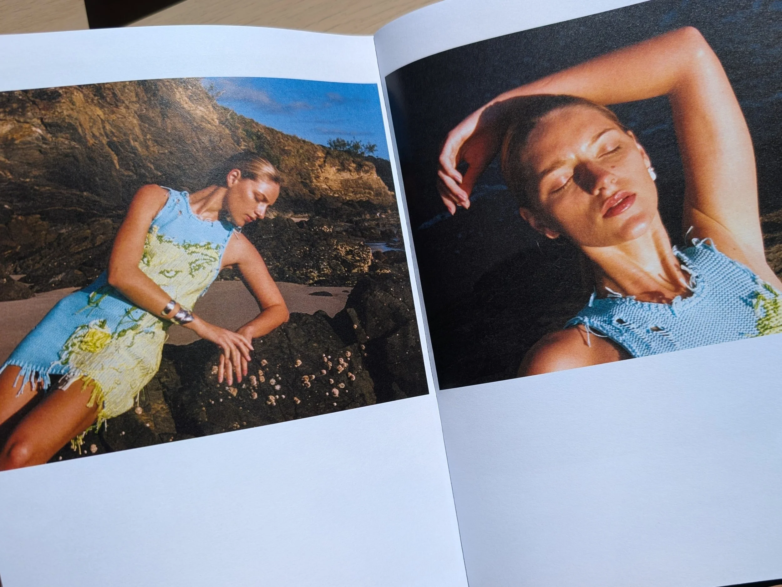 A woman with her eyes closed and arms raised, lying on a rocky beach with cliffs and a blue sky in the background, wearing a sleeveless dress with a floral pattern.