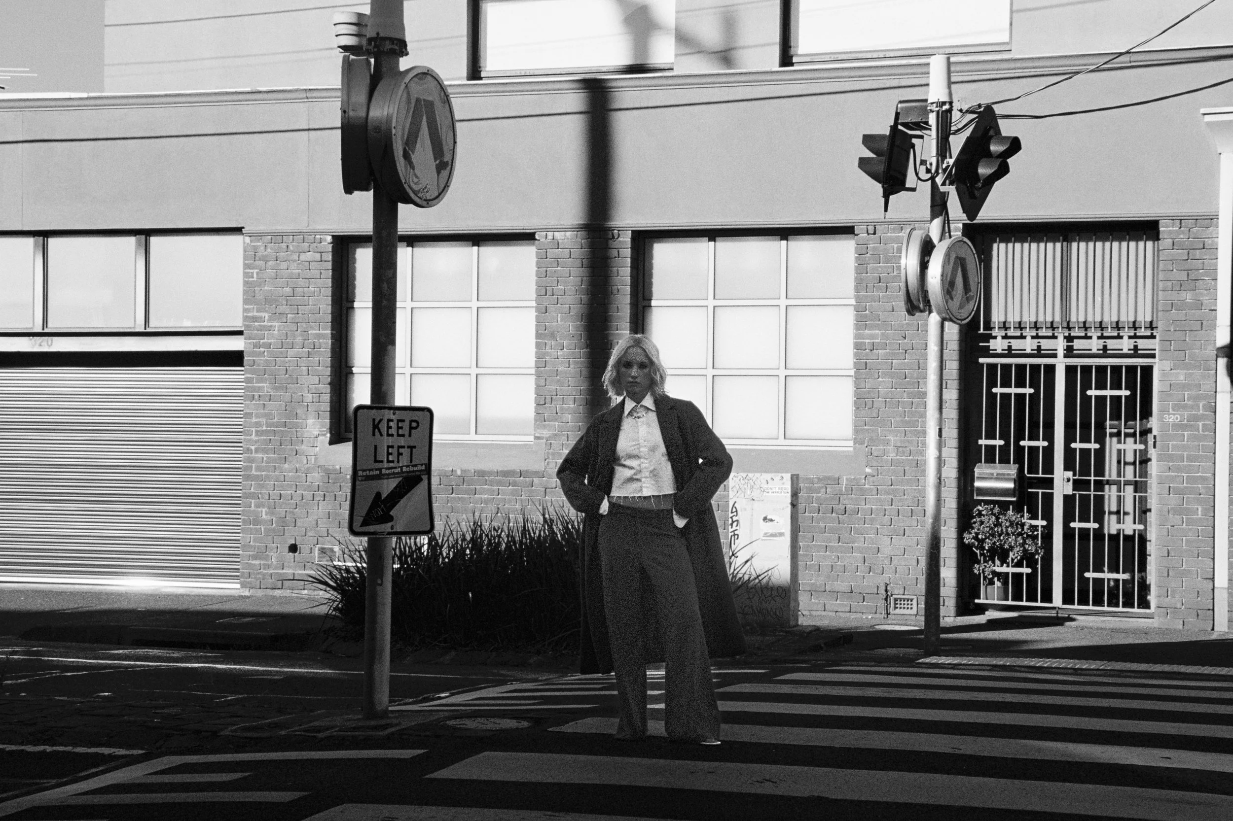 A woman in a blazer and skirt standing at a street corner holding a bag, with traffic lights, street signs, and a building with large windows in the background.