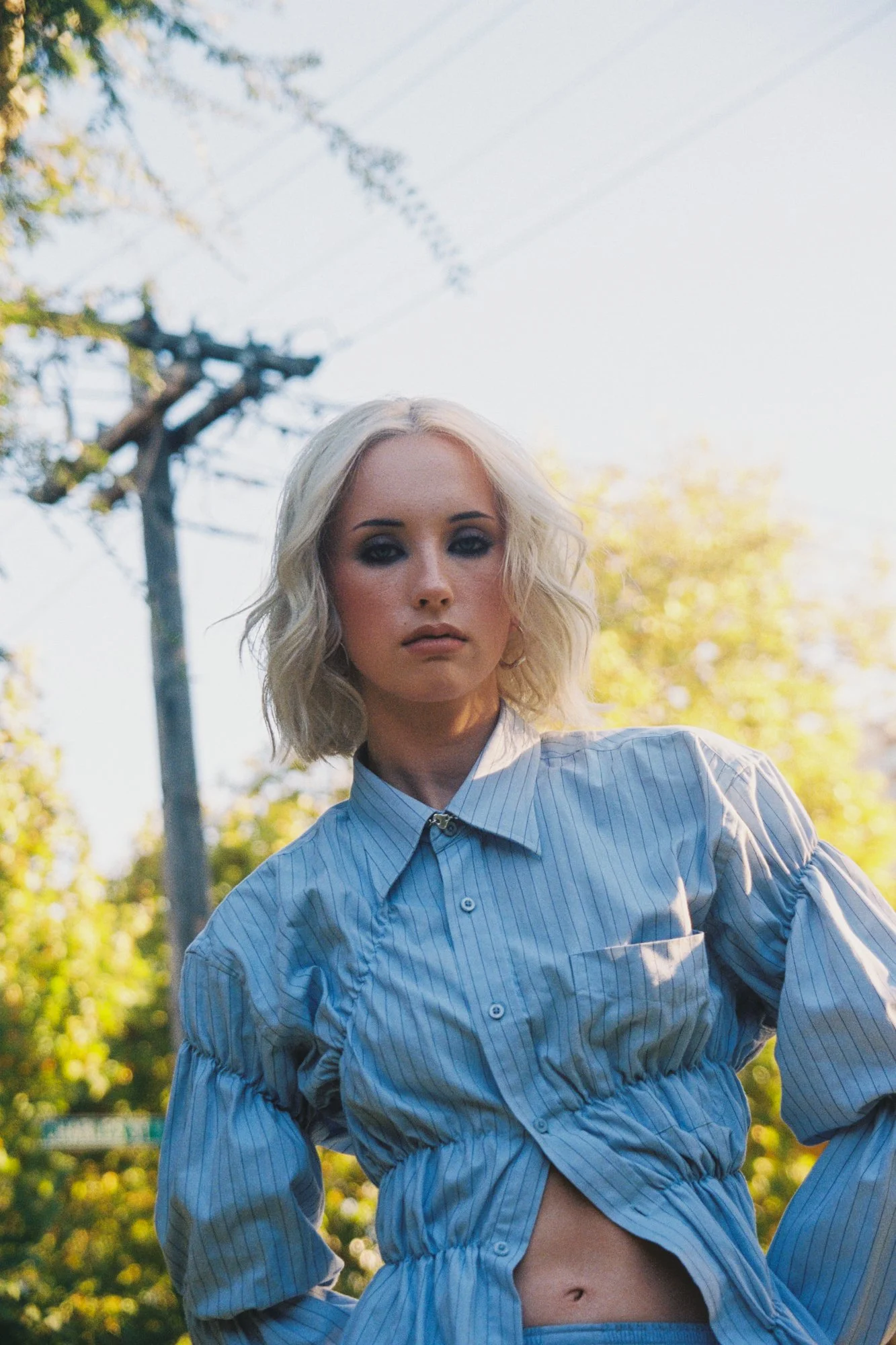 A young woman with platinum blonde hair and blue eyes is standing outdoors in a park during golden hour. She is wearing a blue and white striped button-up shirt that is slightly unbuttoned at the bottom, revealing her midriff.
