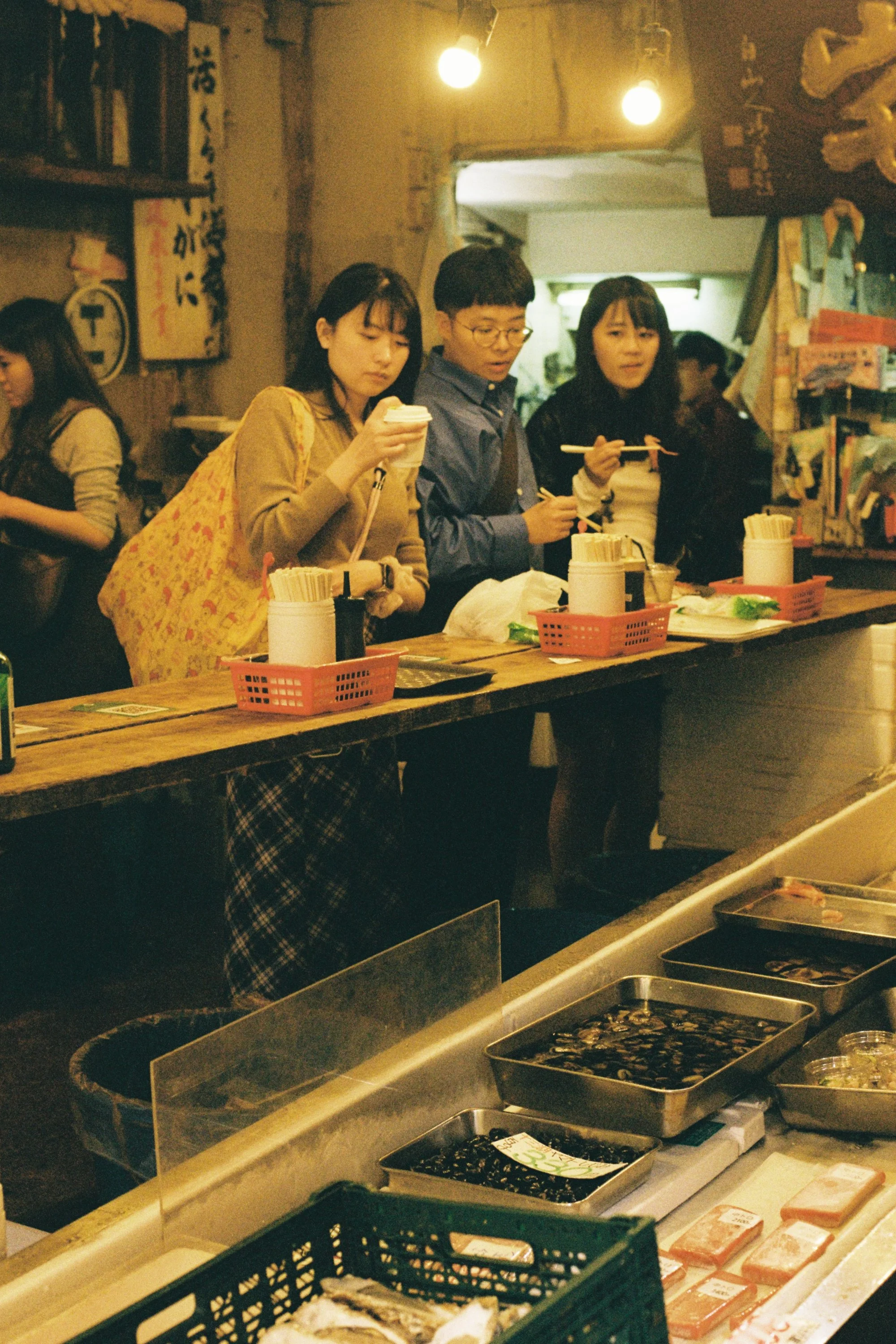 Three people are shopping at a sushi restaurant or market counter, including two women and one man. They are selecting sushi or sashimi using chopsticks. The counter displays various trays of seafood and ingredients, with some of the trays covered in