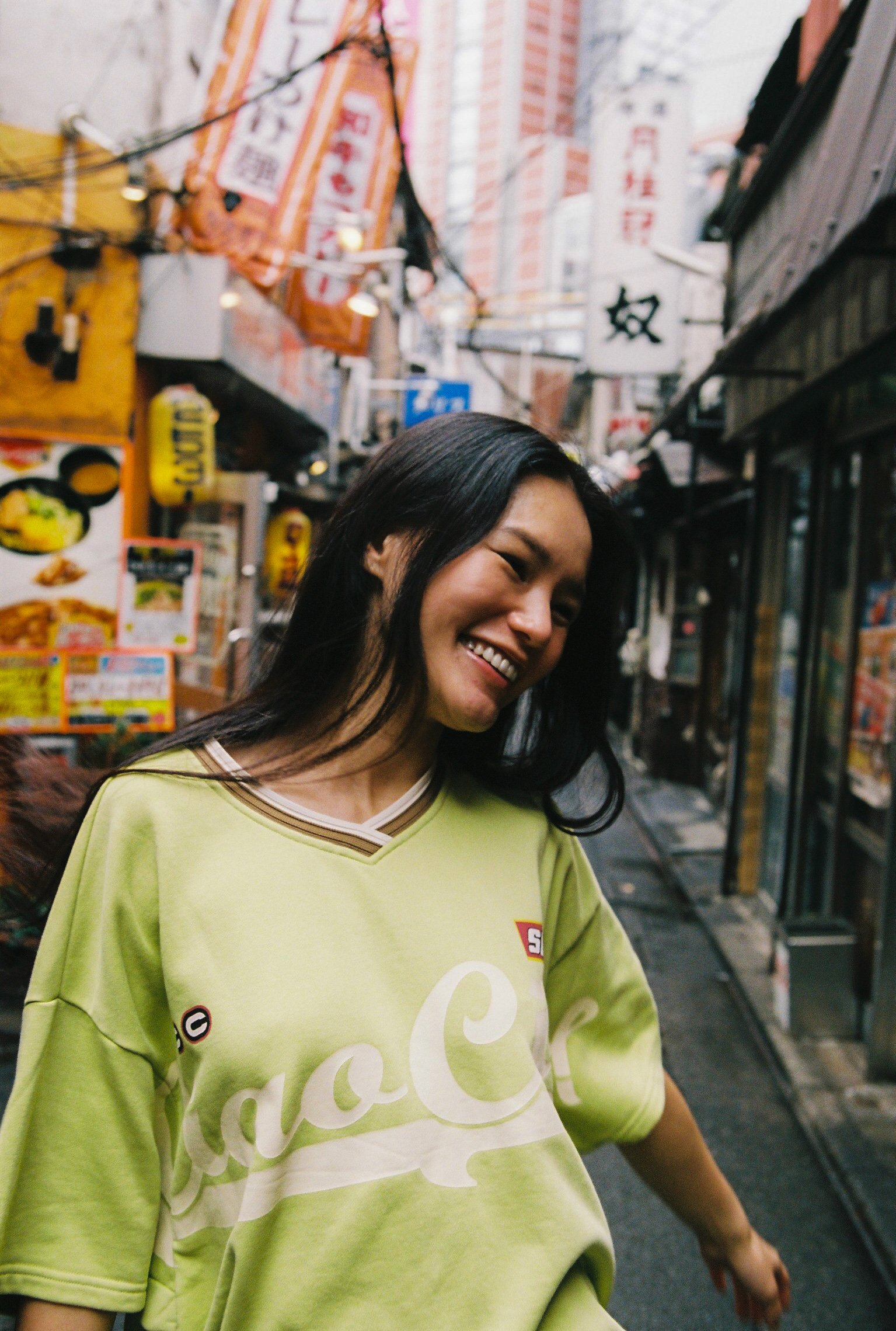 A woman in a neon green oversized t-shirt with a Coca-Cola logo, smiling and walking down a narrow street in an urban area with signs in Chinese characters and hanging lights.