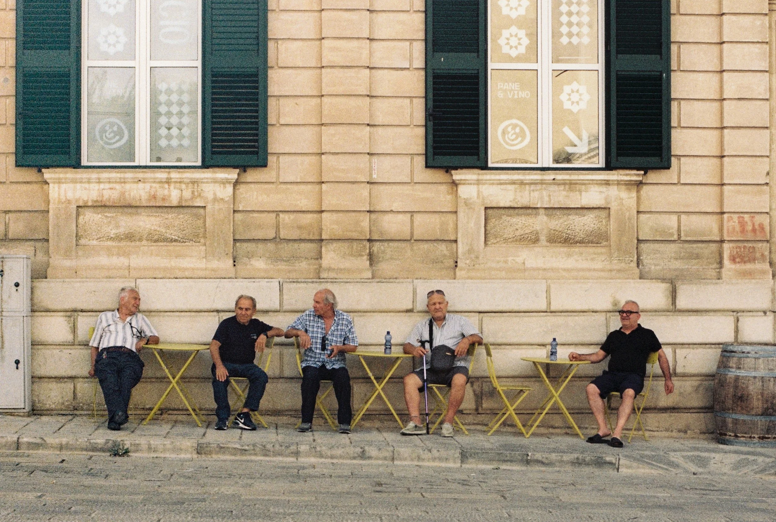 Five older men sitting on yellow chairs outside a building with beige stone walls and dark green shutters, engaged in conversation. There are water bottles on the chairs, and a window with signs indicating a bakery and wine shop in the background.