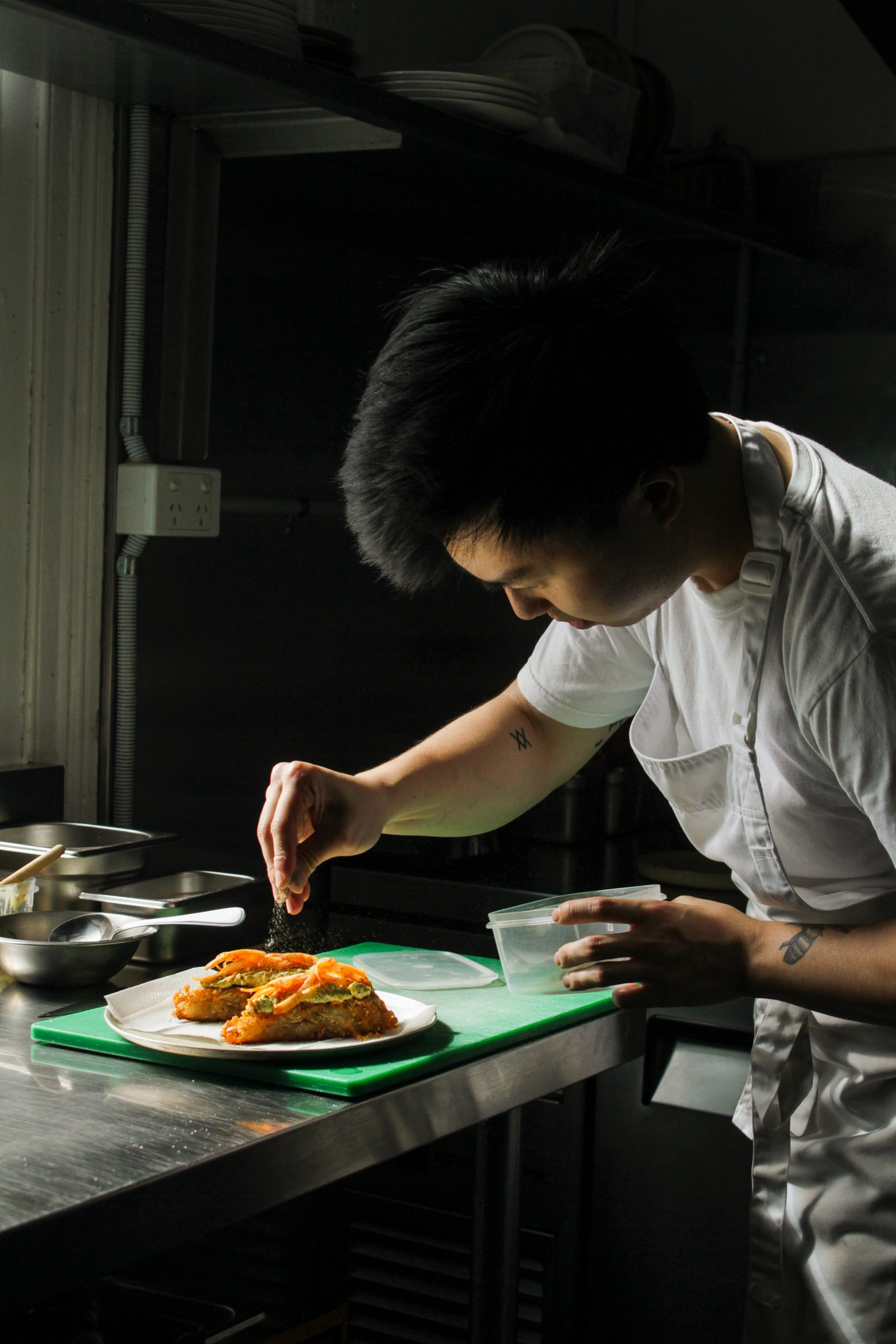 A chef in a white uniform preparing food in a kitchen with dark walls, focusing on plating a dish on a green cutting board. The dish appears to be a seafood or vegetable patty topped with chopped herbs and a drizzle of sauce.