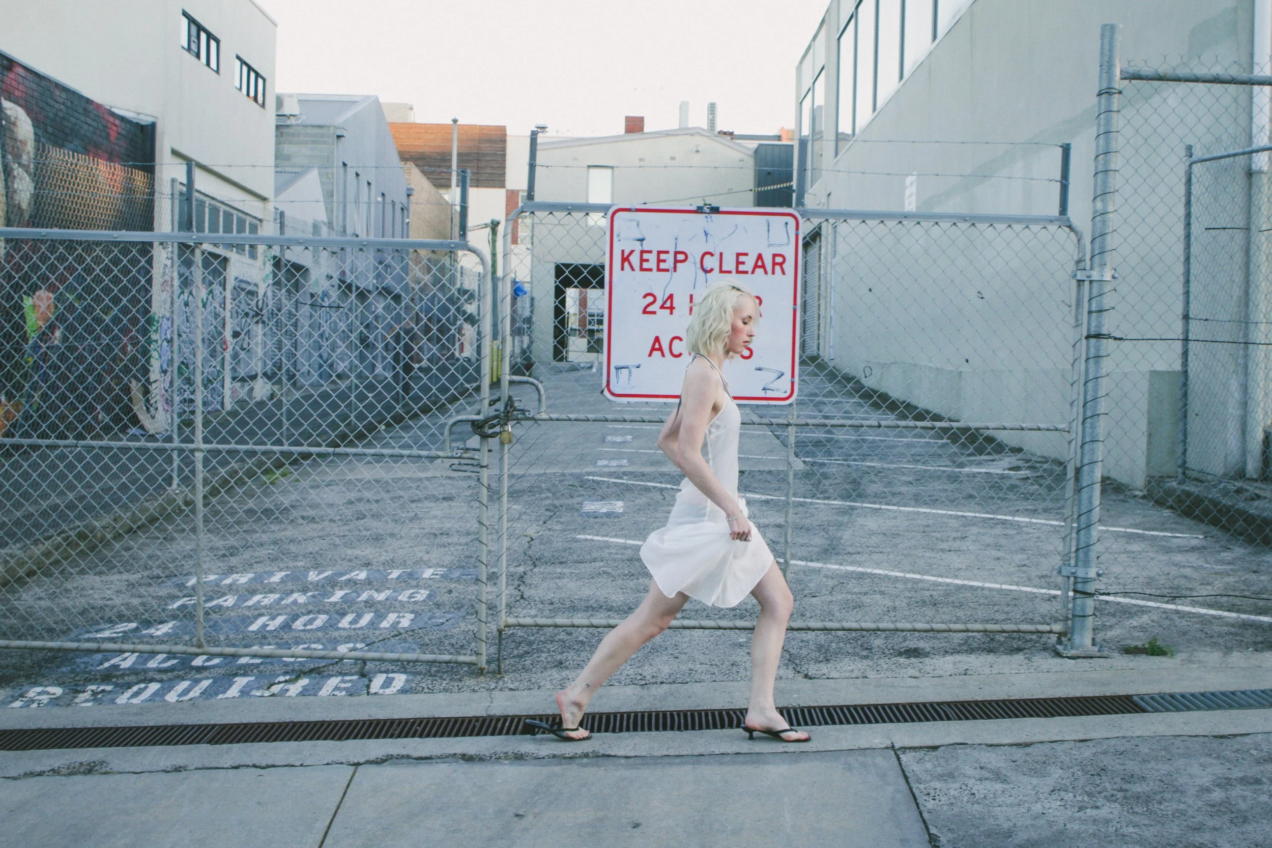 A woman with platinum blonde hair in a white dress and black flip-flops walking past a chain-link gated area with a 'Keep Clear' sign in front of an alleyway in an urban setting.