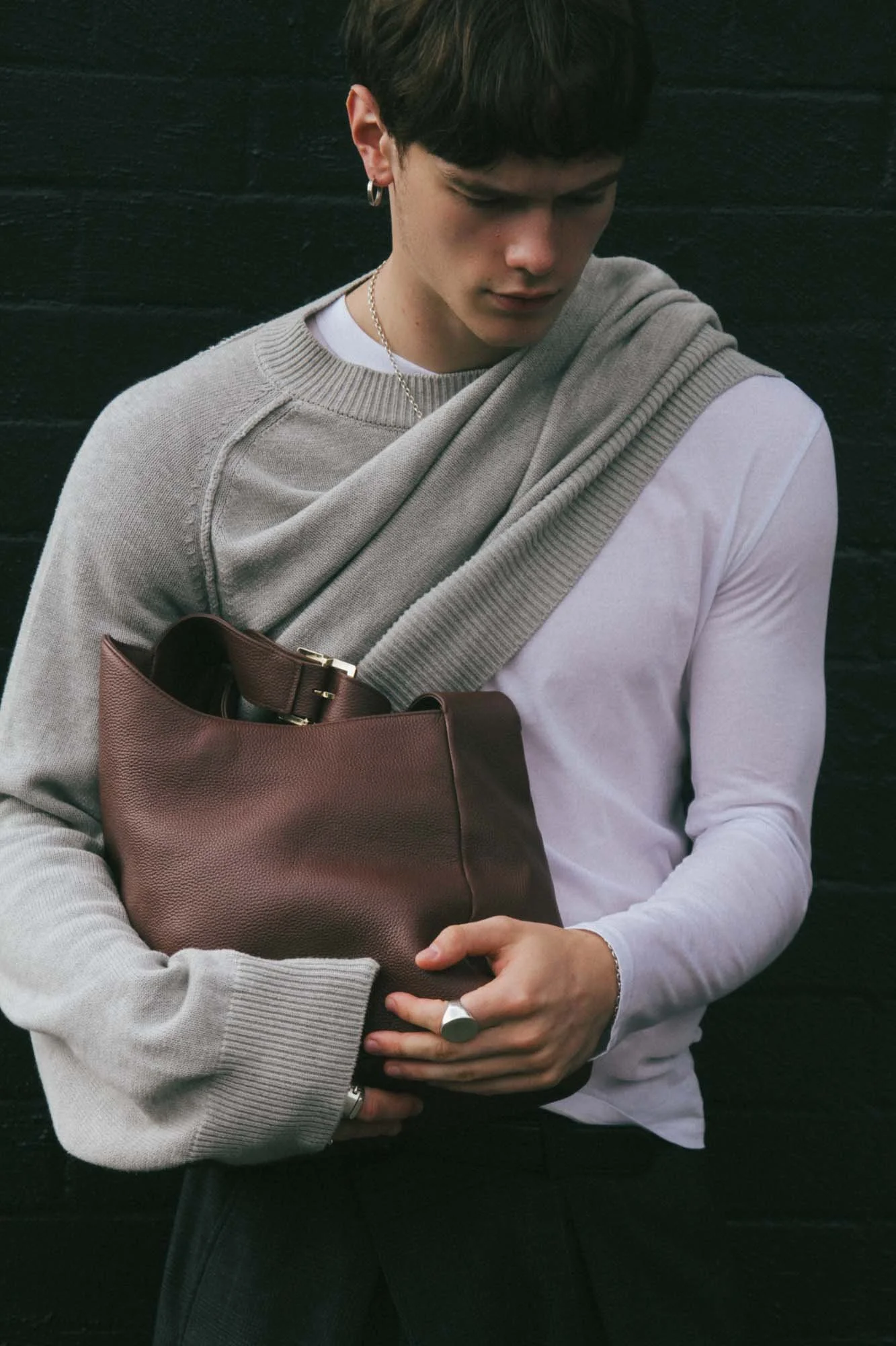 A young man wearing a gray sweater draped over his shoulders, holding a brown handbag, standing against a black wall.