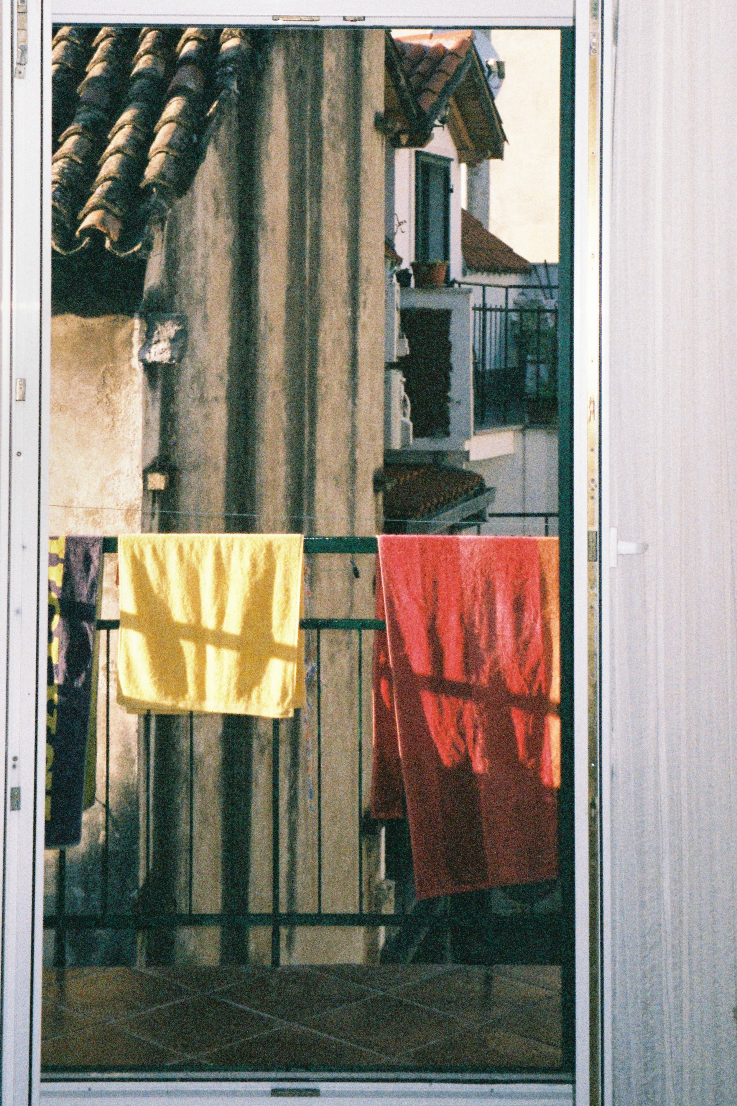 A balcony with clothes hanging to dry, including a yellow towel and a red towel, with a building and a railing visible in the background.