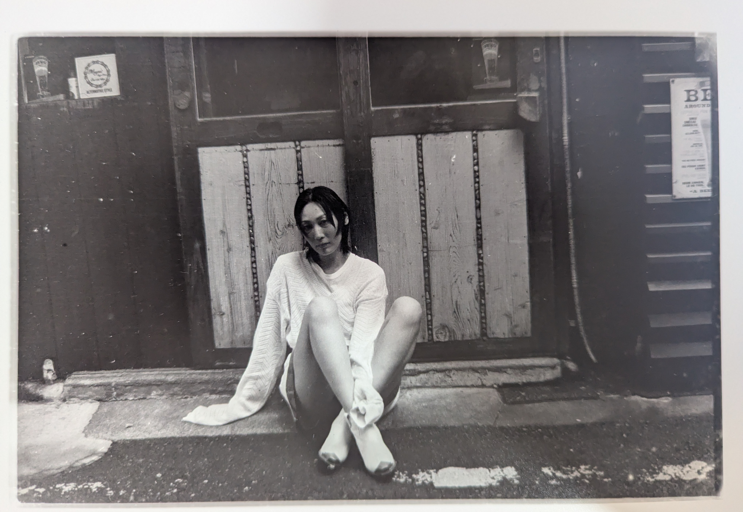 Black and white photo of a young woman sitting on the ground in front of a wooden wall, wearing a white sweater and pants, with her legs extended and crossed at the ankles.