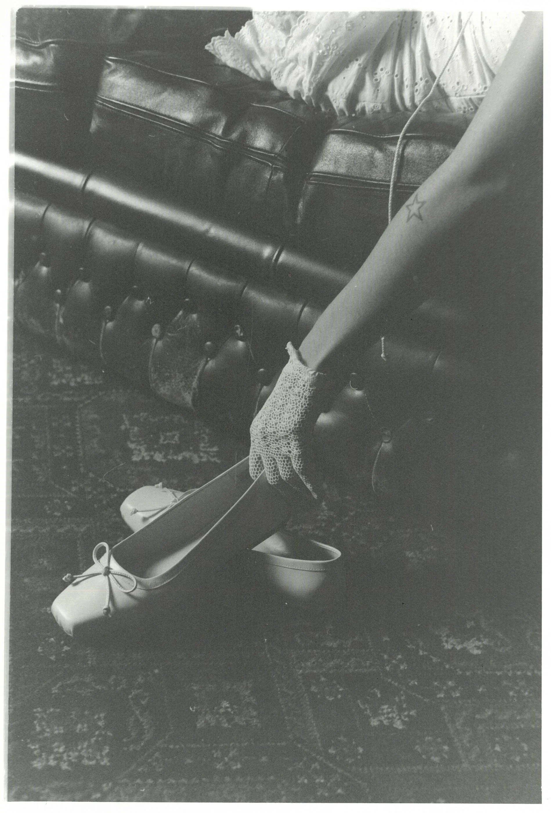 Black and white photograph of a small slide with a metal slide, a car tire, a boots, and a partial view of a person wearing a skirt and a star tattoo on the leg, on a patterned carpet.