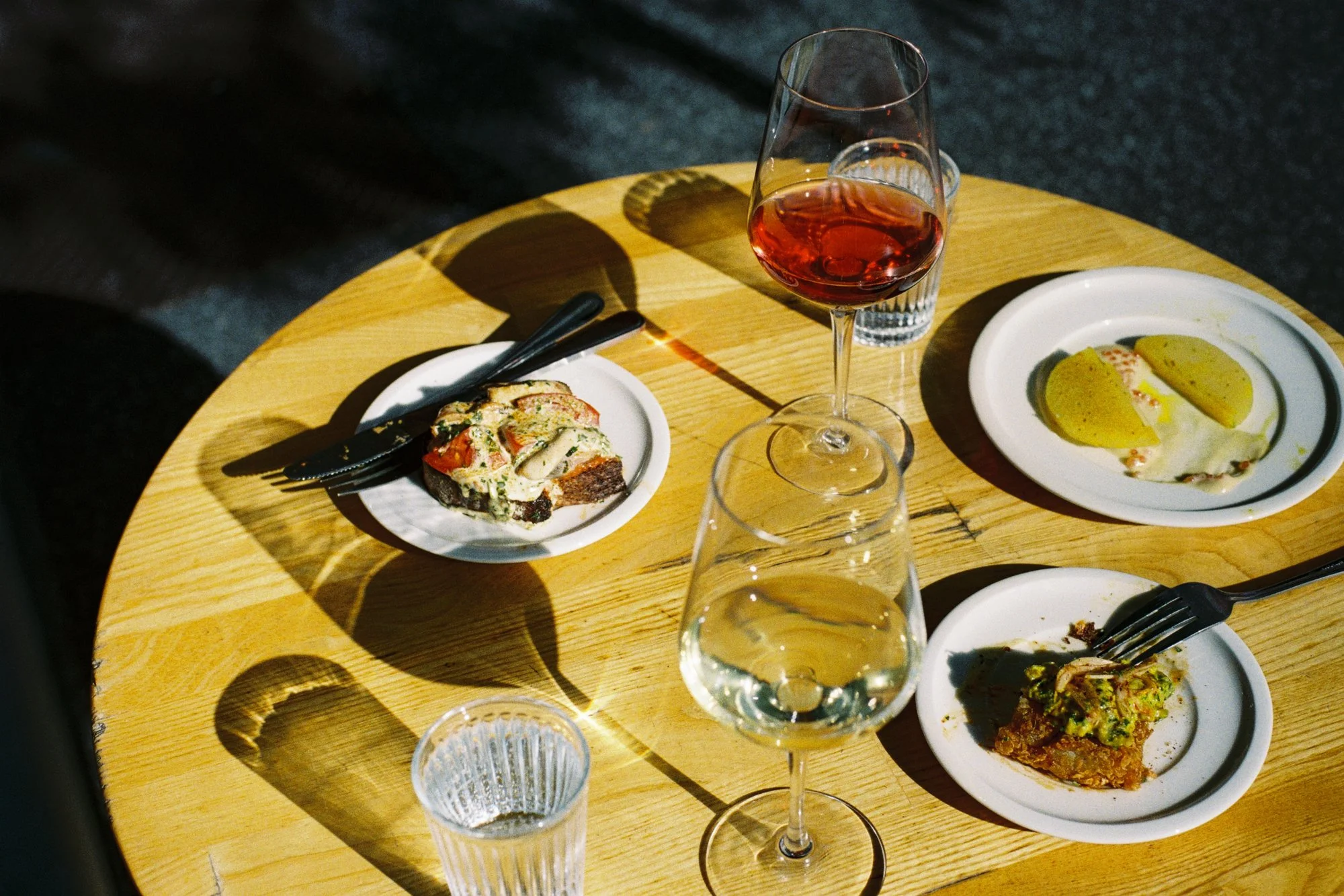 A round wooden table set with a glass of red wine, several empty or partially filled glasses, and small plates with various foods, including a slice of bread topped with cheese and vegetables, and a dish with stuffed peppers, with shadows cast on the