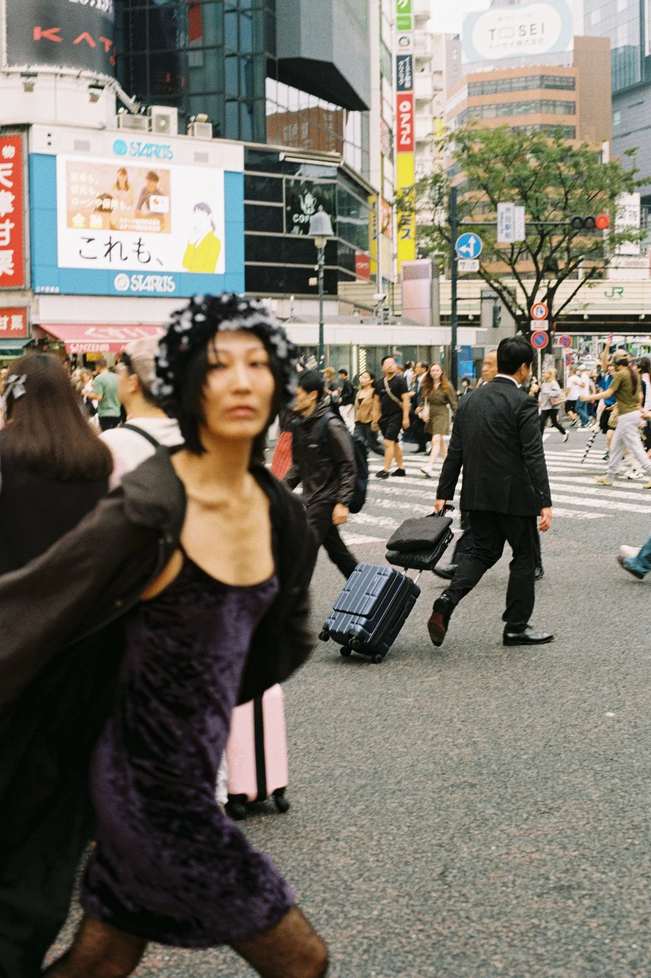 A busy city crosswalk with pedestrians walking, including a woman with a floral headband in the foreground, and numerous buildings with advertisements in the background.