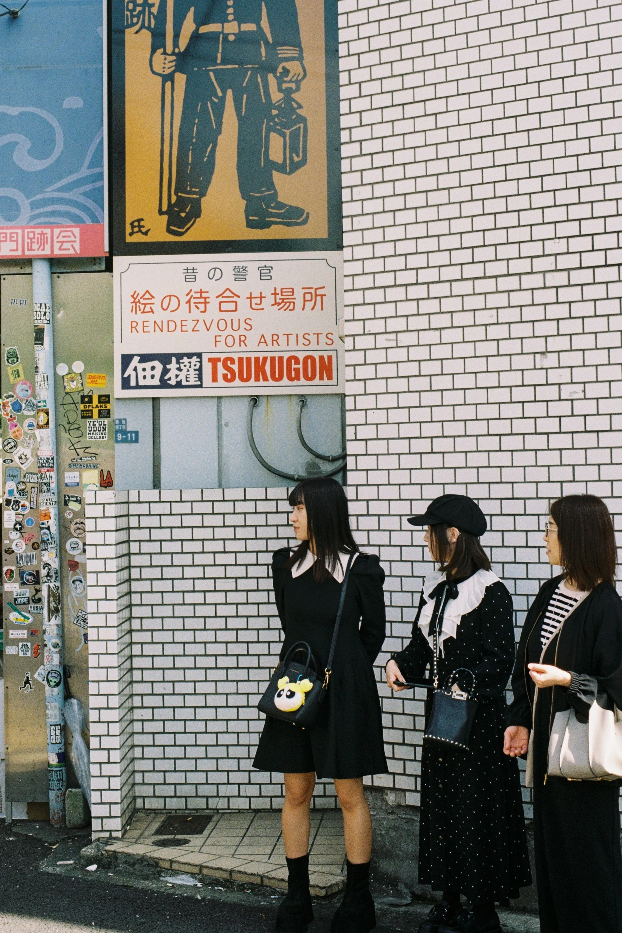 Four women standing in line against a brick wall, dressed in dark clothing, with one woman carrying a black bag with a plush toy, and three women holding their phones.