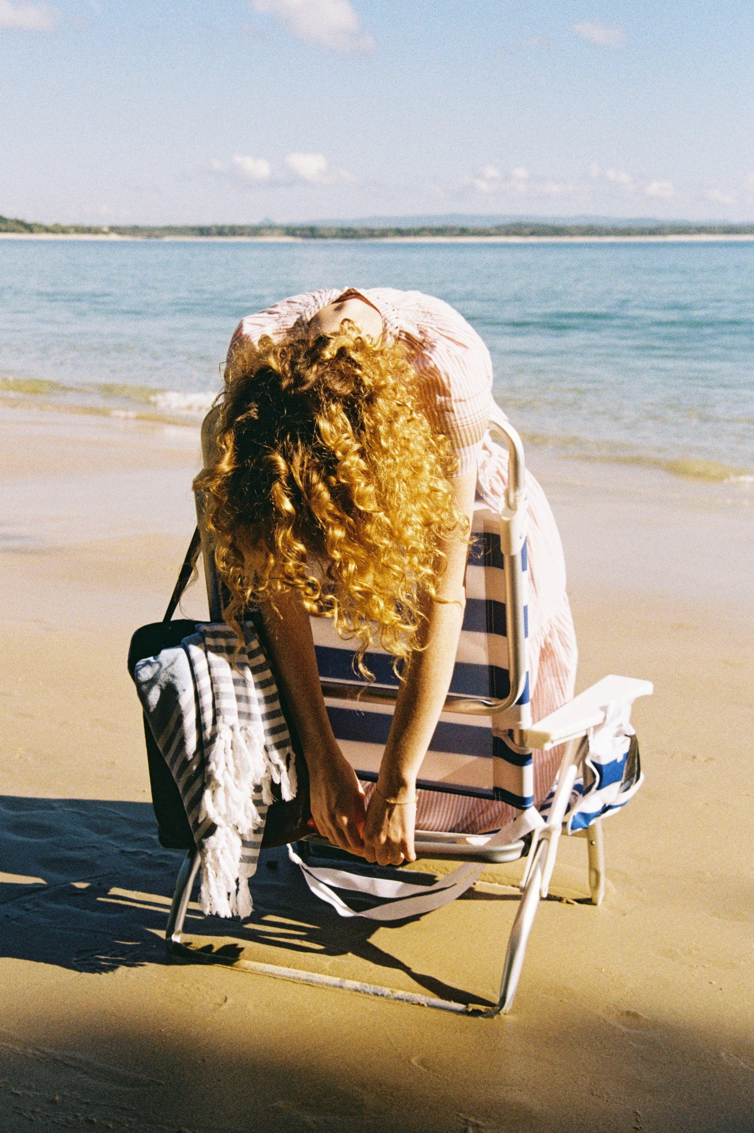 A woman with long curly hair sitting in a beach chair on the sand near the water, with her head tilted back and her arms resting on her lap, wearing a pink and white striped dress. A striped towel hangs from the chair, and a black and white bag is on