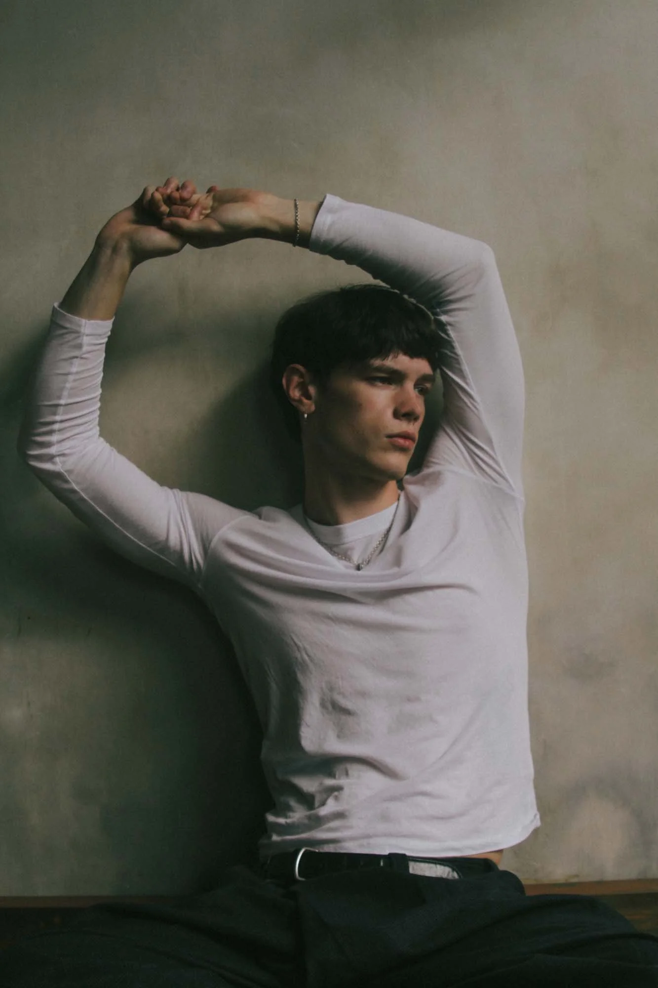 Young man with short dark hair and earrings, wearing a white long-sleeve shirt, laying on a flat surface with arms raised above his head, against a neutral wall background.
