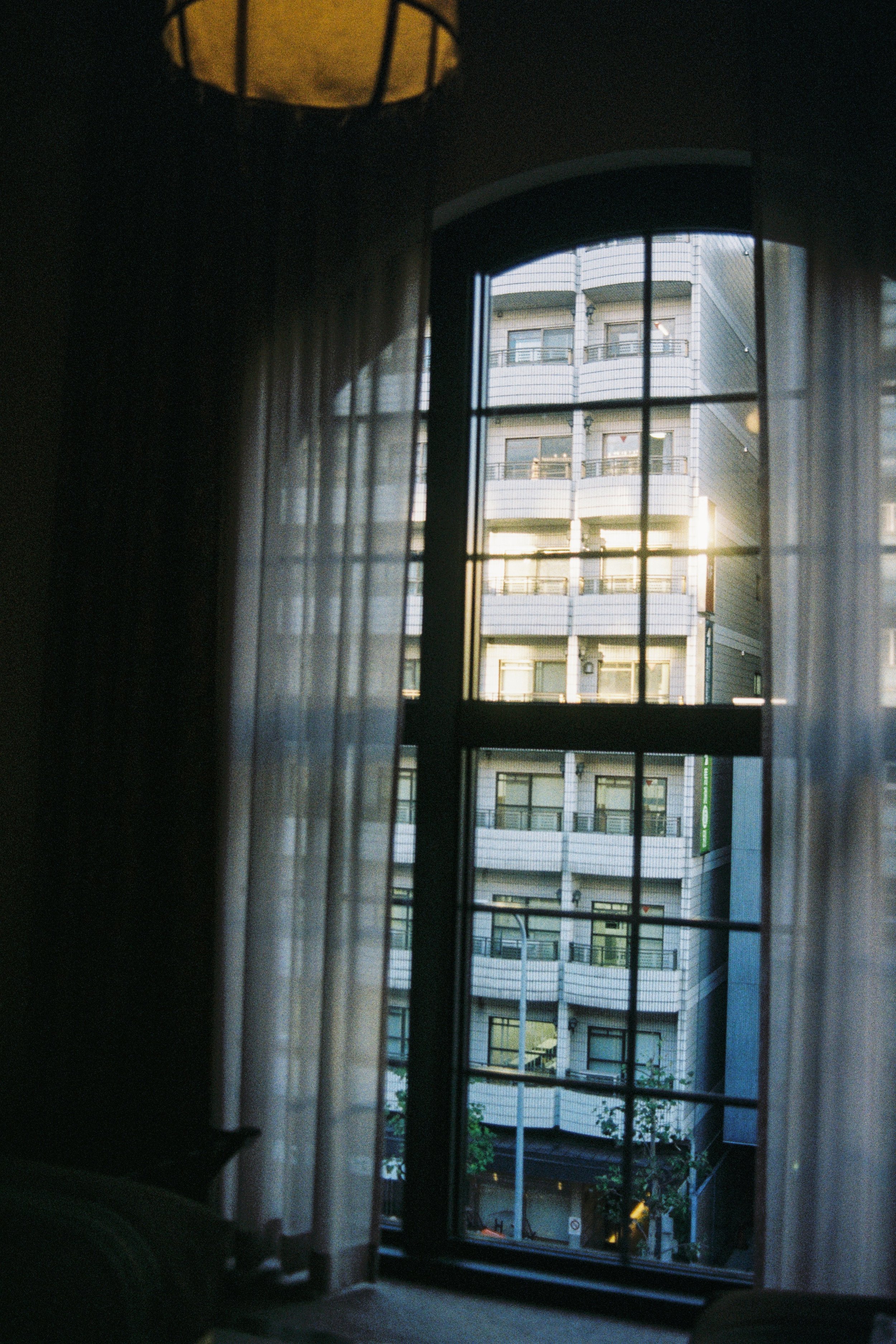 View of a high-rise apartment building through a window with sheer curtains, sunlight illuminating the building at sunset.