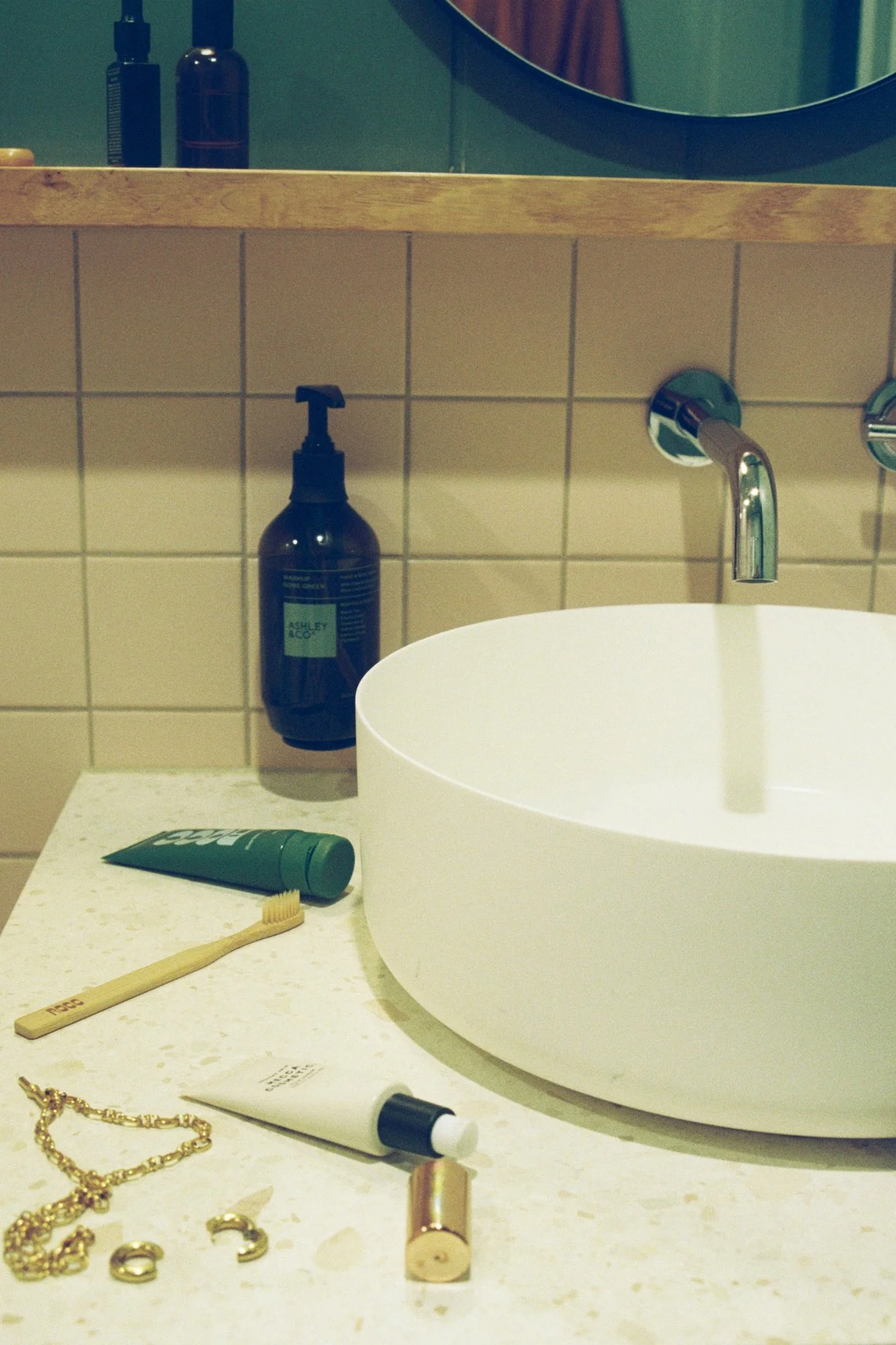 Bathroom countertop with jewelry, a toothbrush, toothpaste, and various toiletries including a soap dispenser and lotion. A round sink and a mirror are also visible.