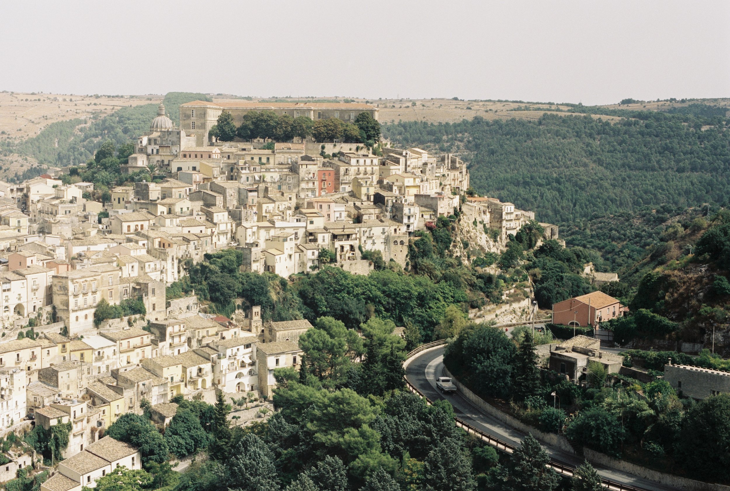 A hillside town with numerous white and beige buildings and a large historic fort or castle on top, surrounded by green trees and open countryside, with a winding road and a vehicle in the foreground.