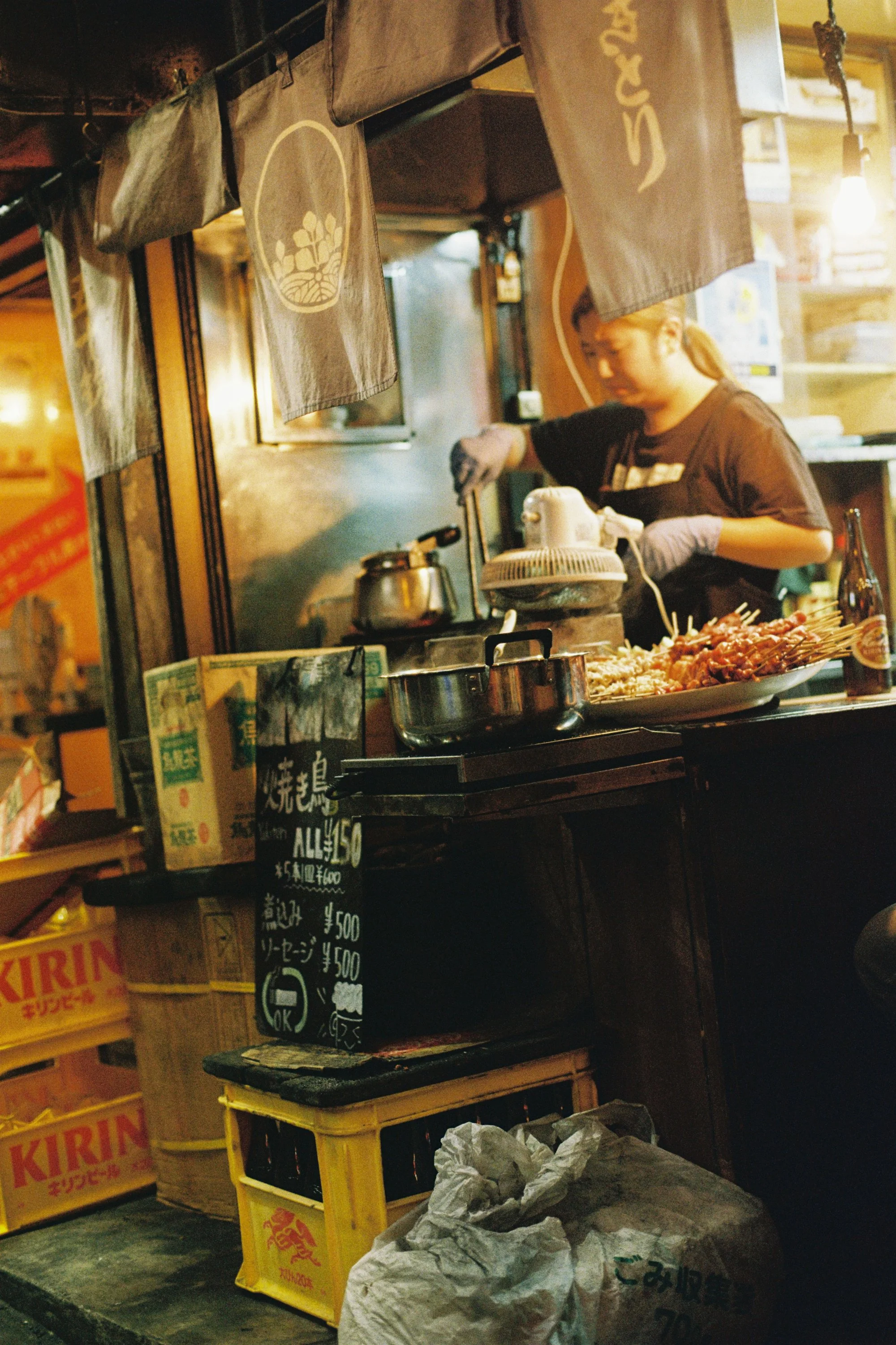 A woman is working behind a food stall with various food items and Japanese signs and prices displayed. There are cardboard boxes and paper bags at the front.