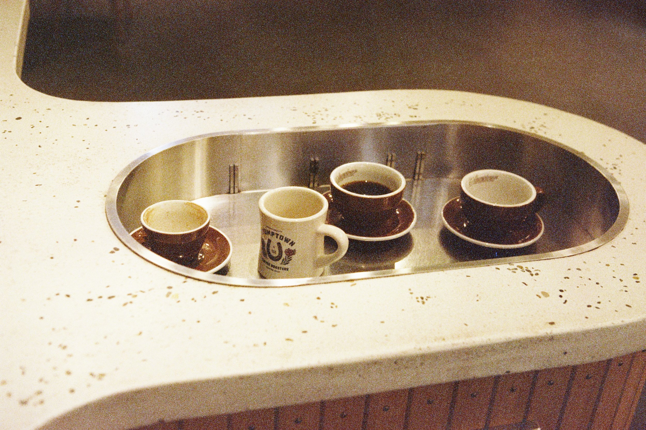 Six coffee cups on a round tray inside a kitchen counter sink, with one white mug having printed text and a graphic.