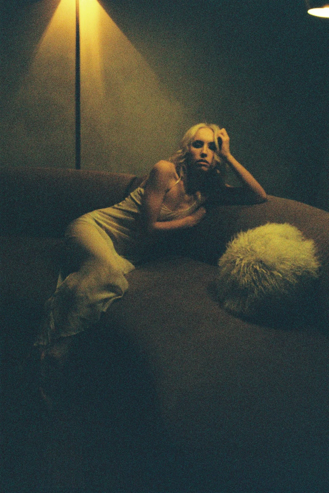 A woman with blonde hair reclining on a dark couch in a dimly lit room, with a white fluffy dog resting beside her.