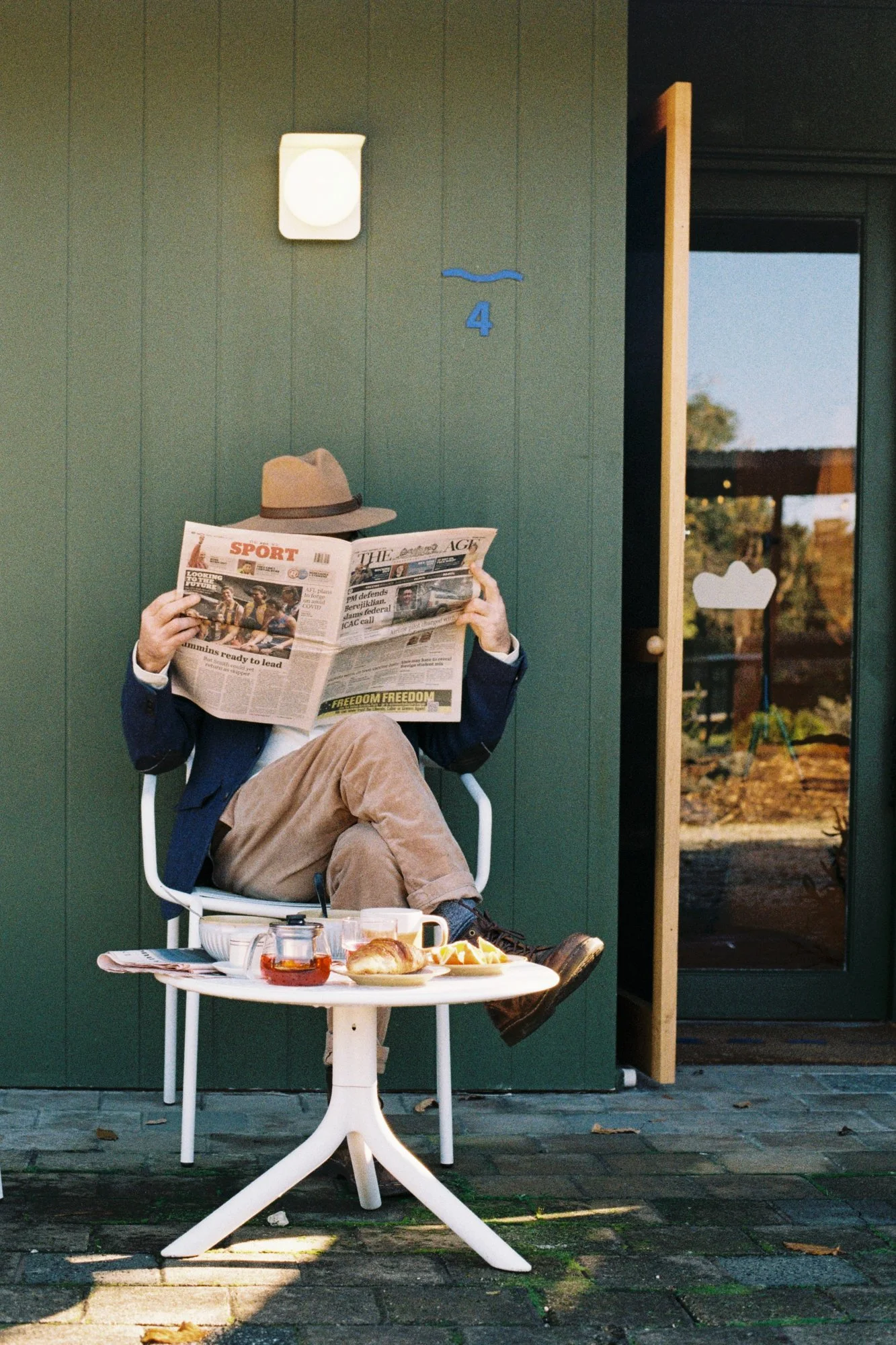 Person sitting outside at a small round table with breakfast, reading a newspaper, wearing a fedora, blazer, and khakis, with a green building and blue sky in the background.