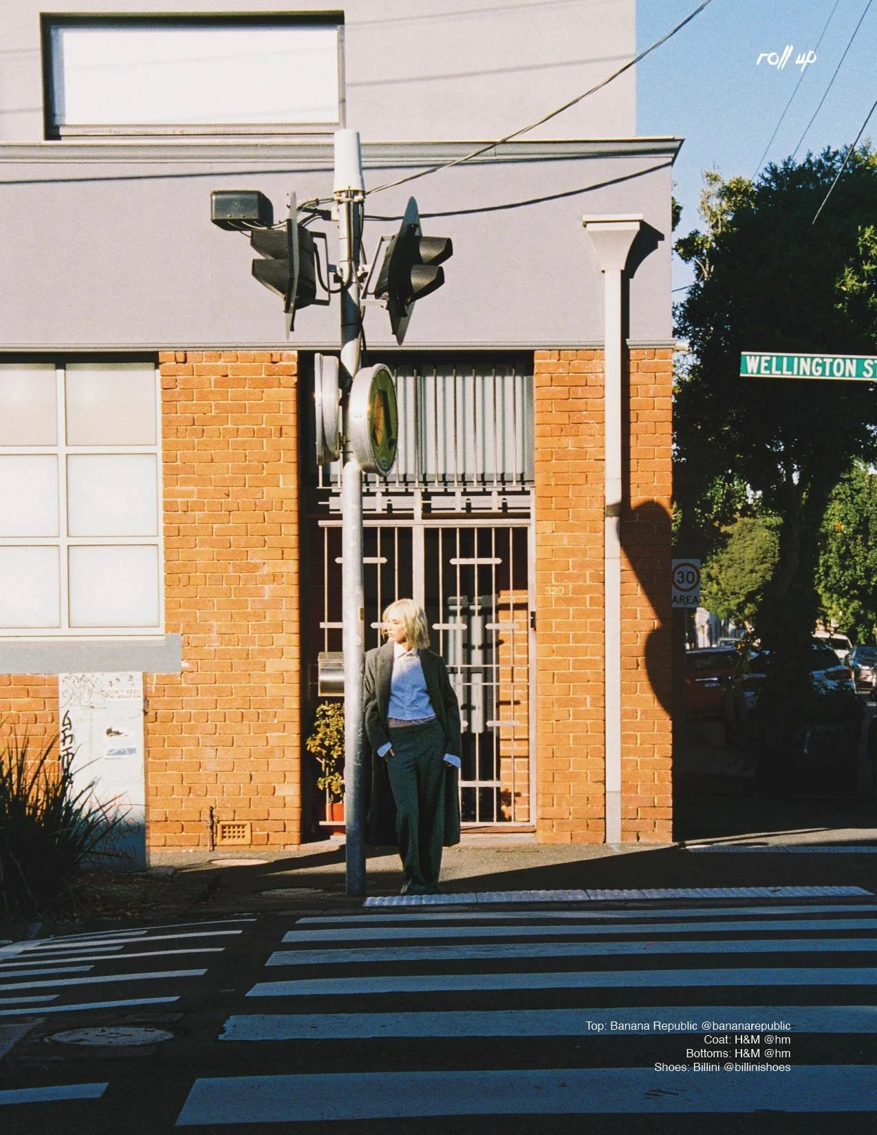 A woman with blond hair wearing a dark coat, white shirt, blue pants, and black shoes standing at a crosswalk on a city street corner. She is standing in front of a red brick building with a barred door and a window. There is a street sign reading "W