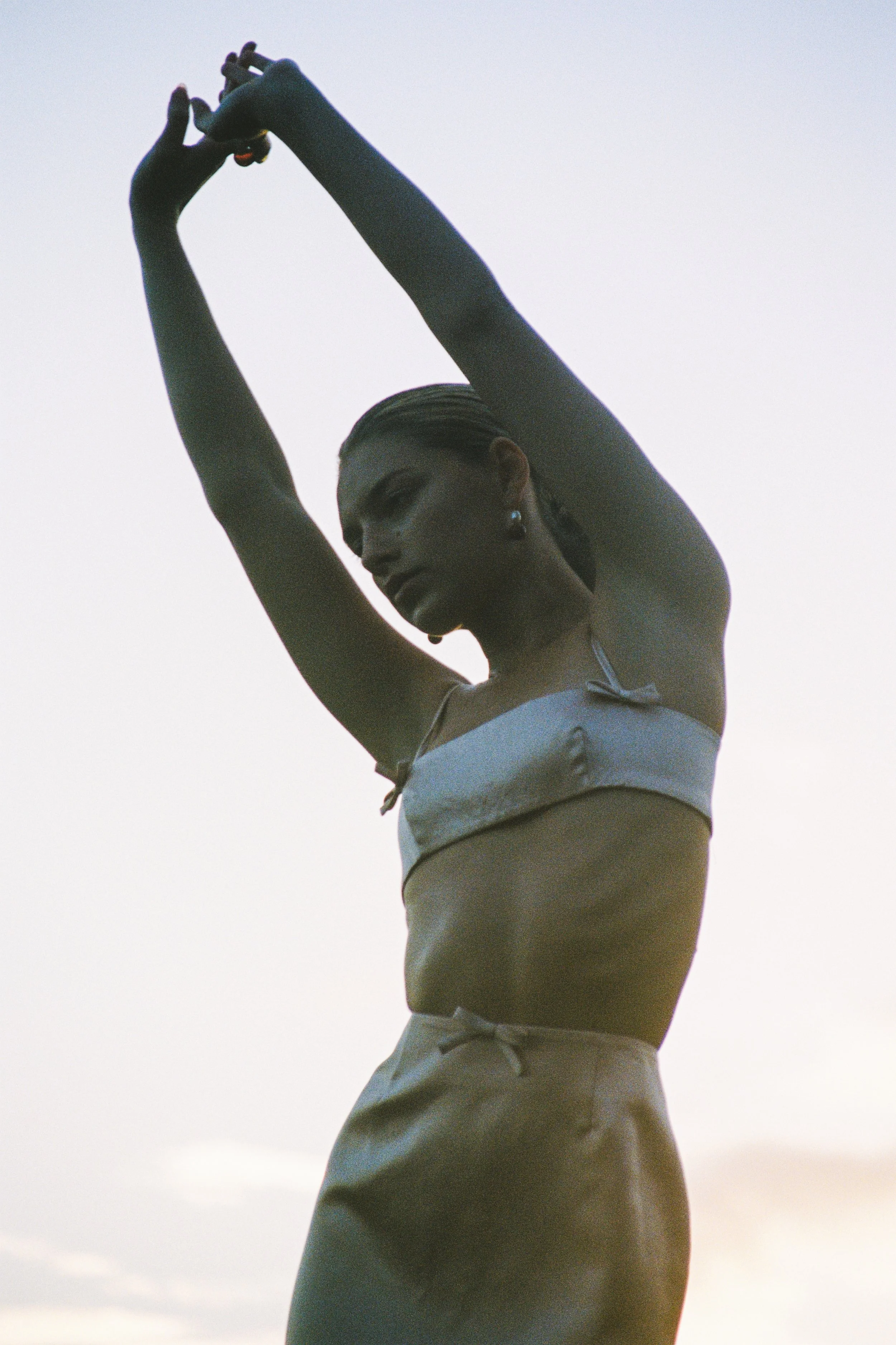 Close-up of a woman with braided hair, earrings, minimal makeup, wearing a white tank top and beige shorts, standing outdoors against a cloudy sky, with her hands raised above her head.