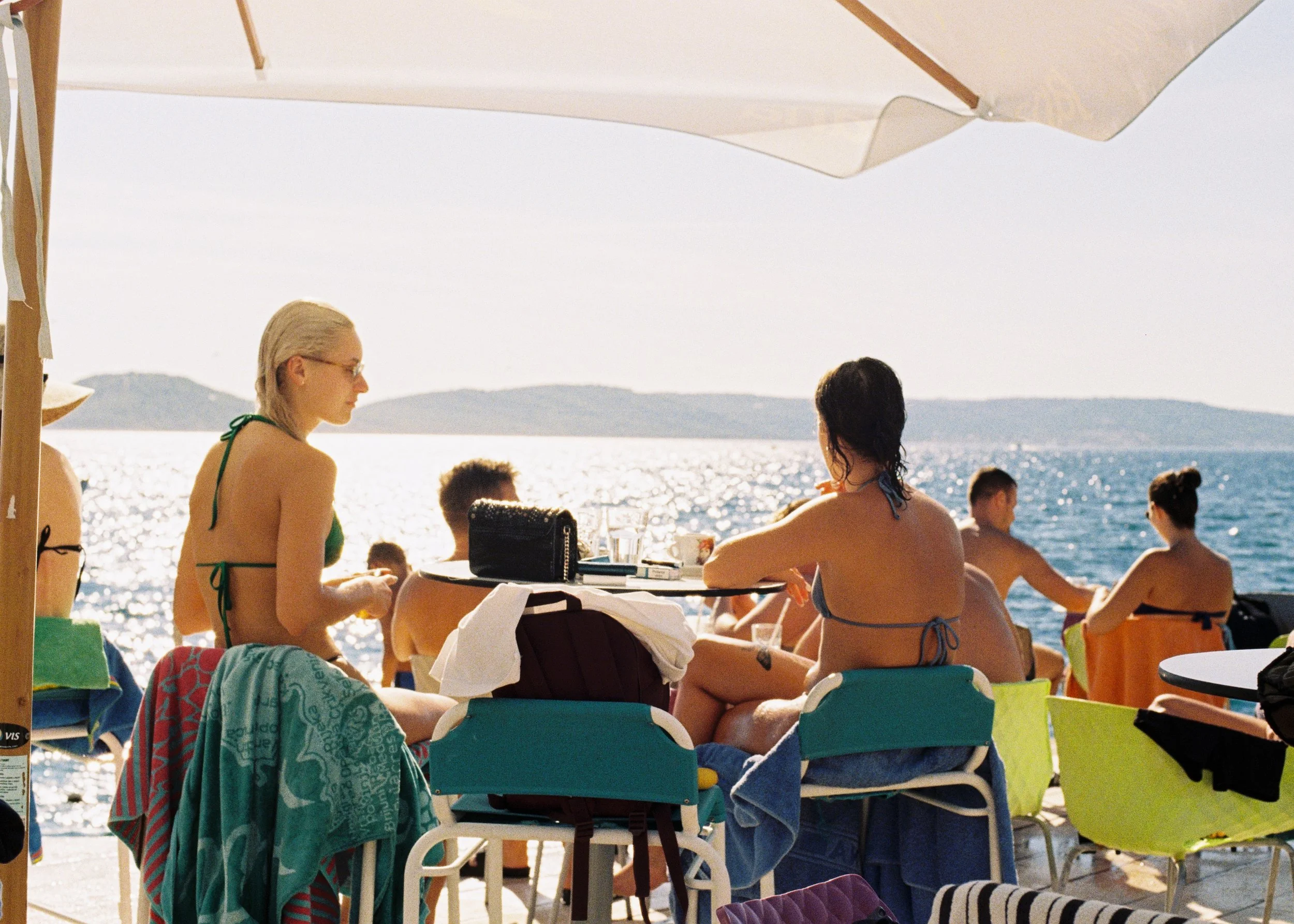 People sitting and relaxing at an outdoor beachside café with a view of the sea and distant islands, some wearing swimsuits and sunglasses, under large umbrellas.