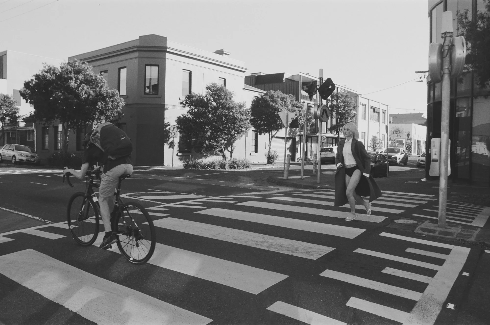 A woman in a long coat and high heels crossing a city street at a crosswalk as a cyclist rides by. Buildings, trees, cars, and street signs are visible in the background, along with traffic lights and a bus stop.