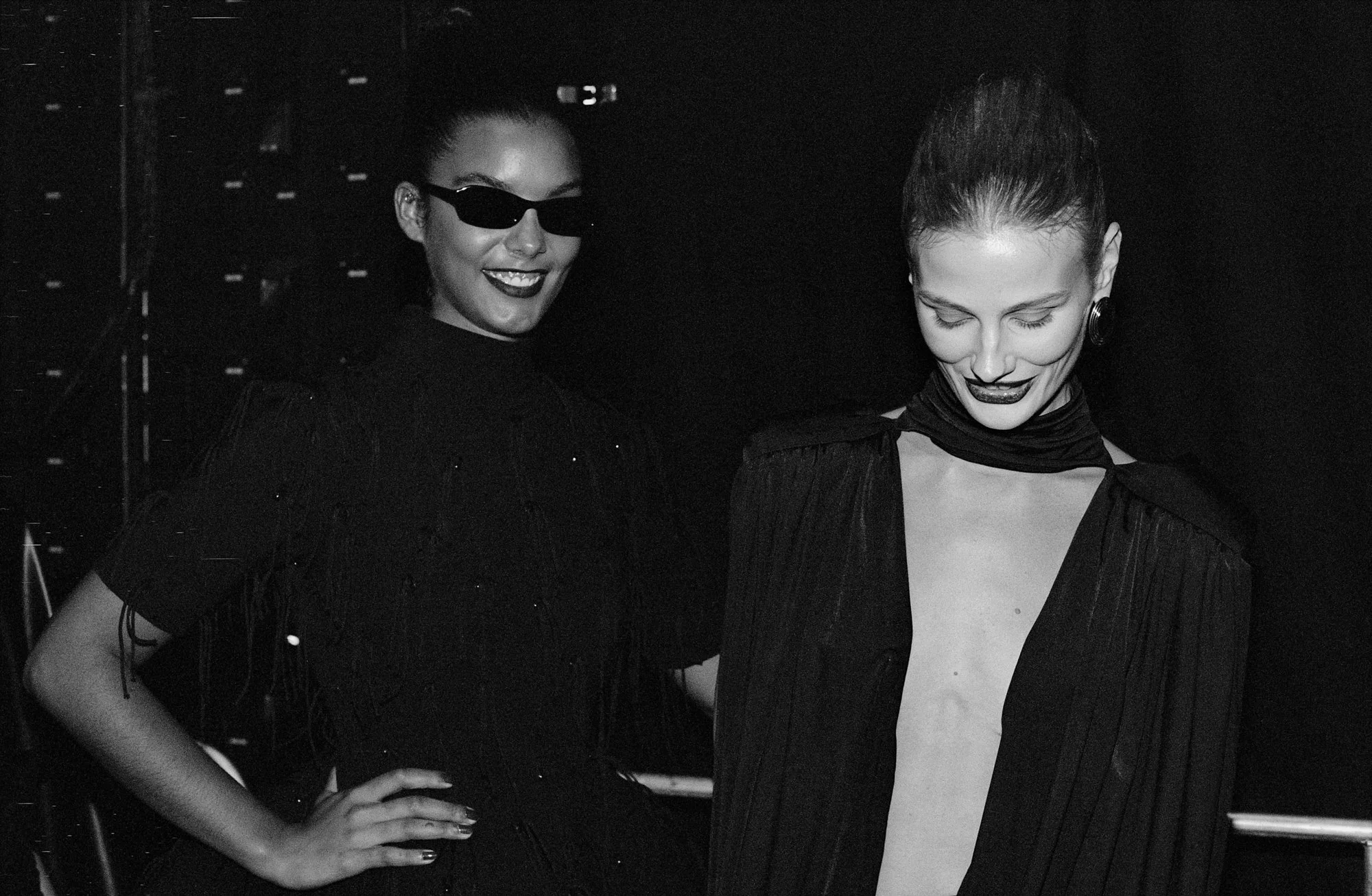 Two women dressed in dark clothing, one wearing sunglasses, smiling and posing for the camera in a dark setting.