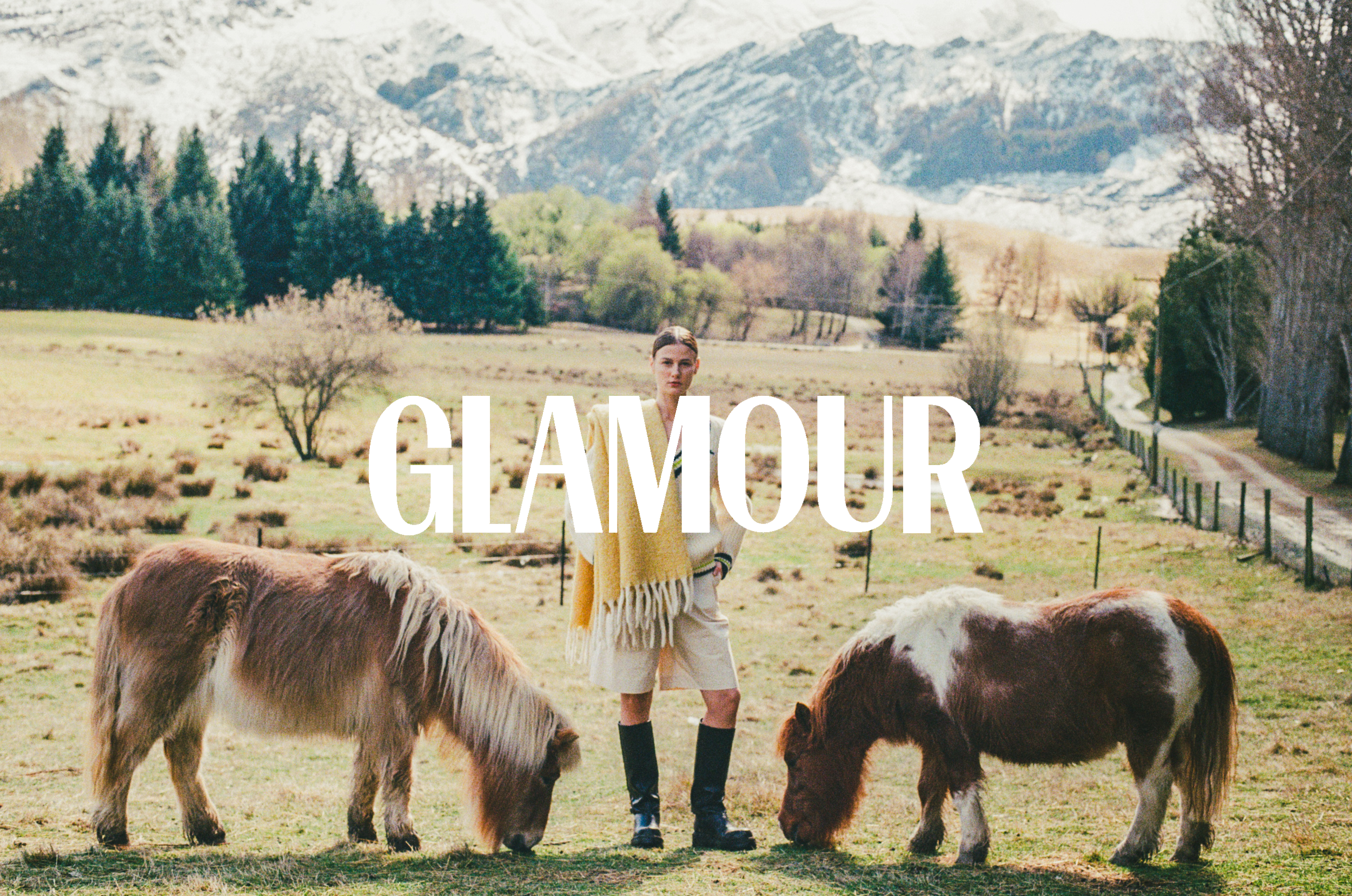 A woman standing among several cows on a farm with a mountainous background, and the word 'GLAMOUR' written across the image.