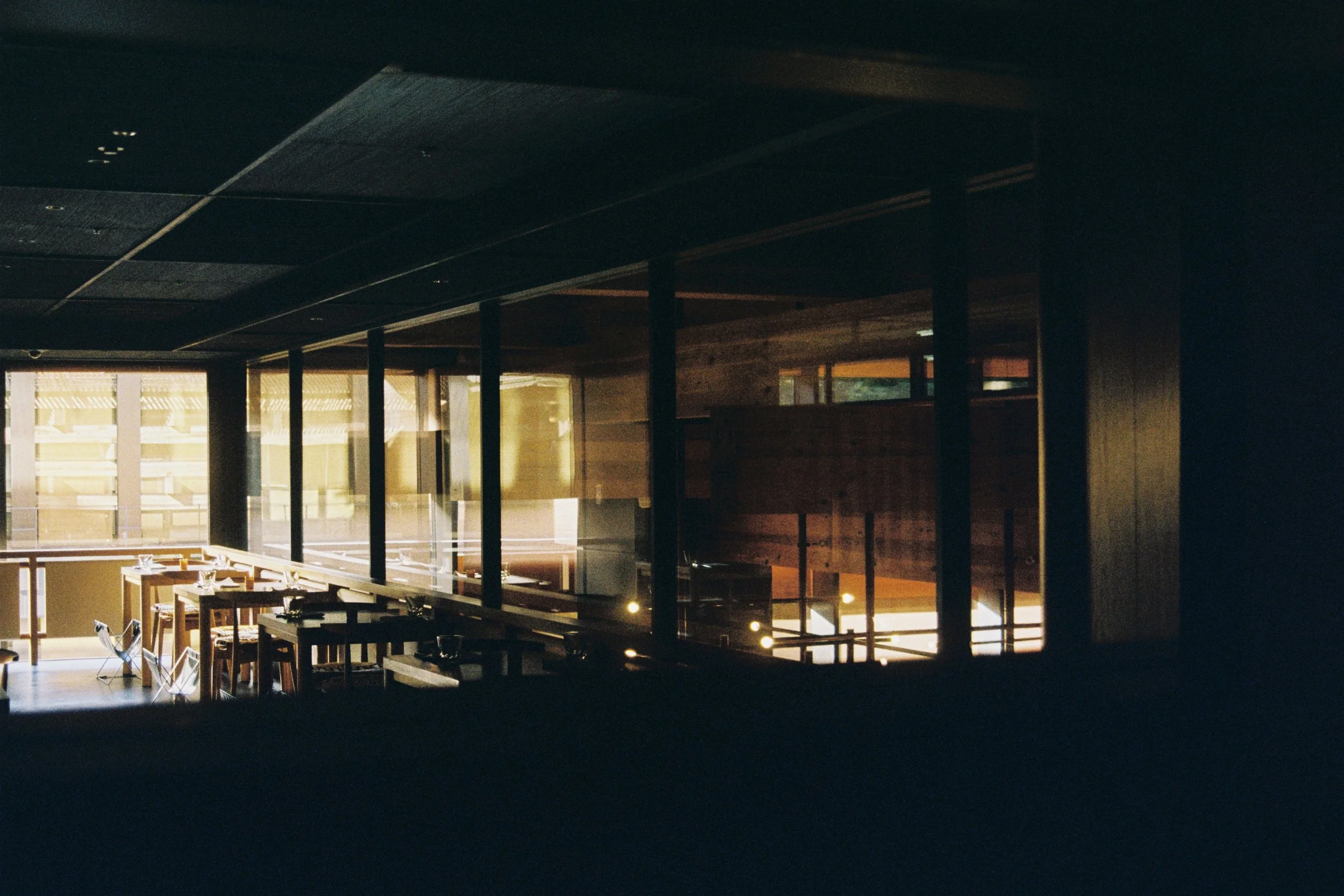 Interior of a modern restaurant with wooden furniture and large windows, illuminated by warm lighting.