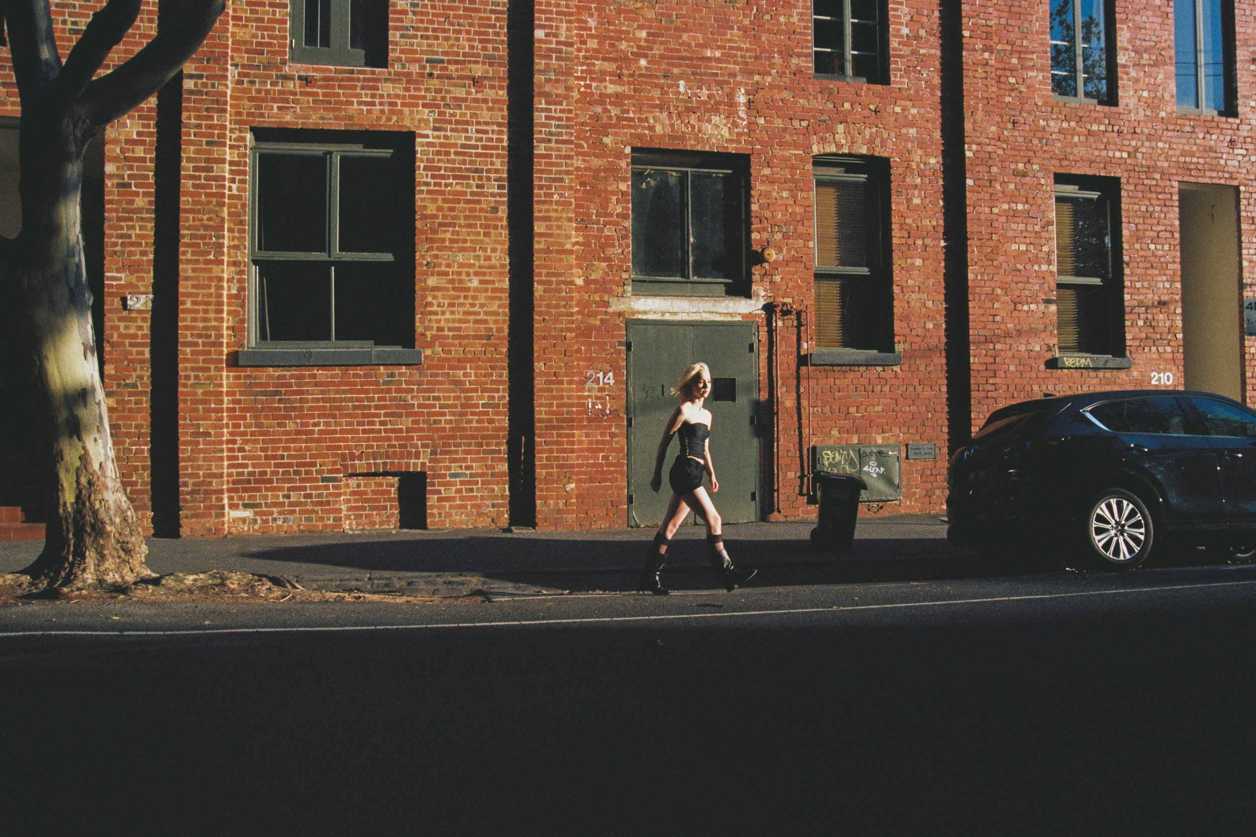 A woman wearing a black strapless top and black shorts walking past a brick building with multiple windows, a door, and a parked black car. A tree with peeling bark is on the left side of the image, and there is graffiti on a small structure near the