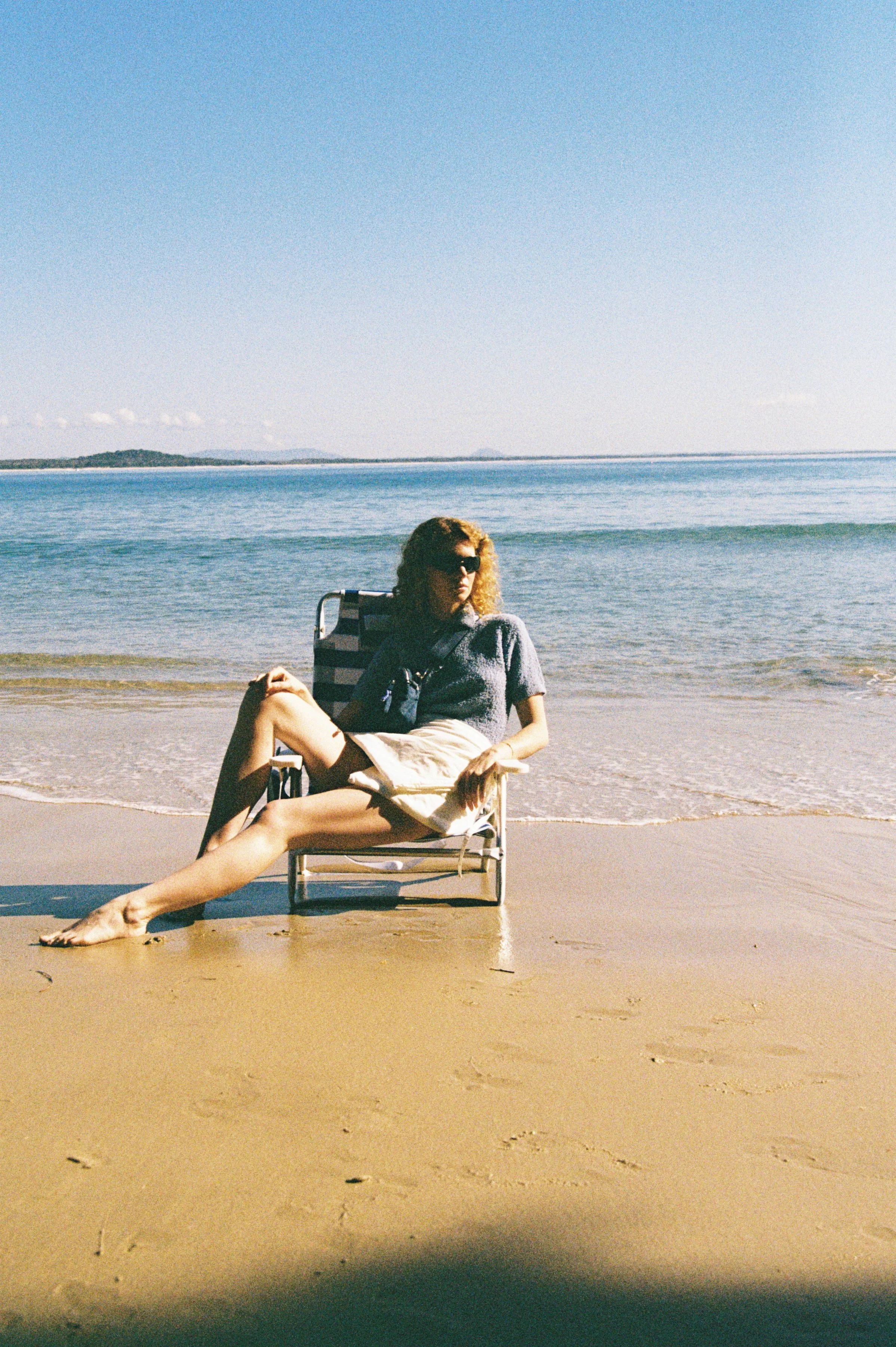 A woman with curly hair and sunglasses relaxing on a beach chair with her legs crossed over the sand. The background features the ocean with gentle waves and a distant shoreline under a clear sky.