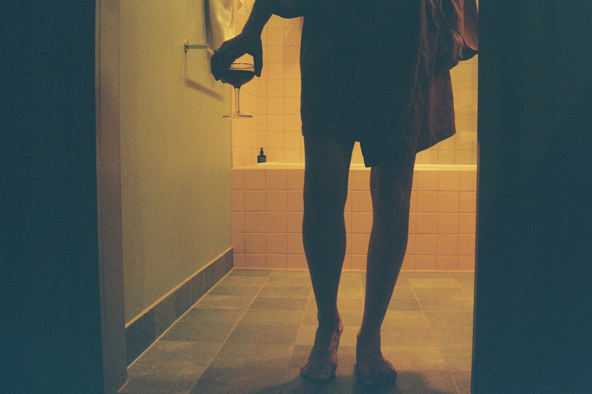 A person taking a mirror selfie in a dimly lit bathroom, holding a wine glass with dark liquid. The person's face is not visible, wearing dark clothing and standing in front of tiled walls.