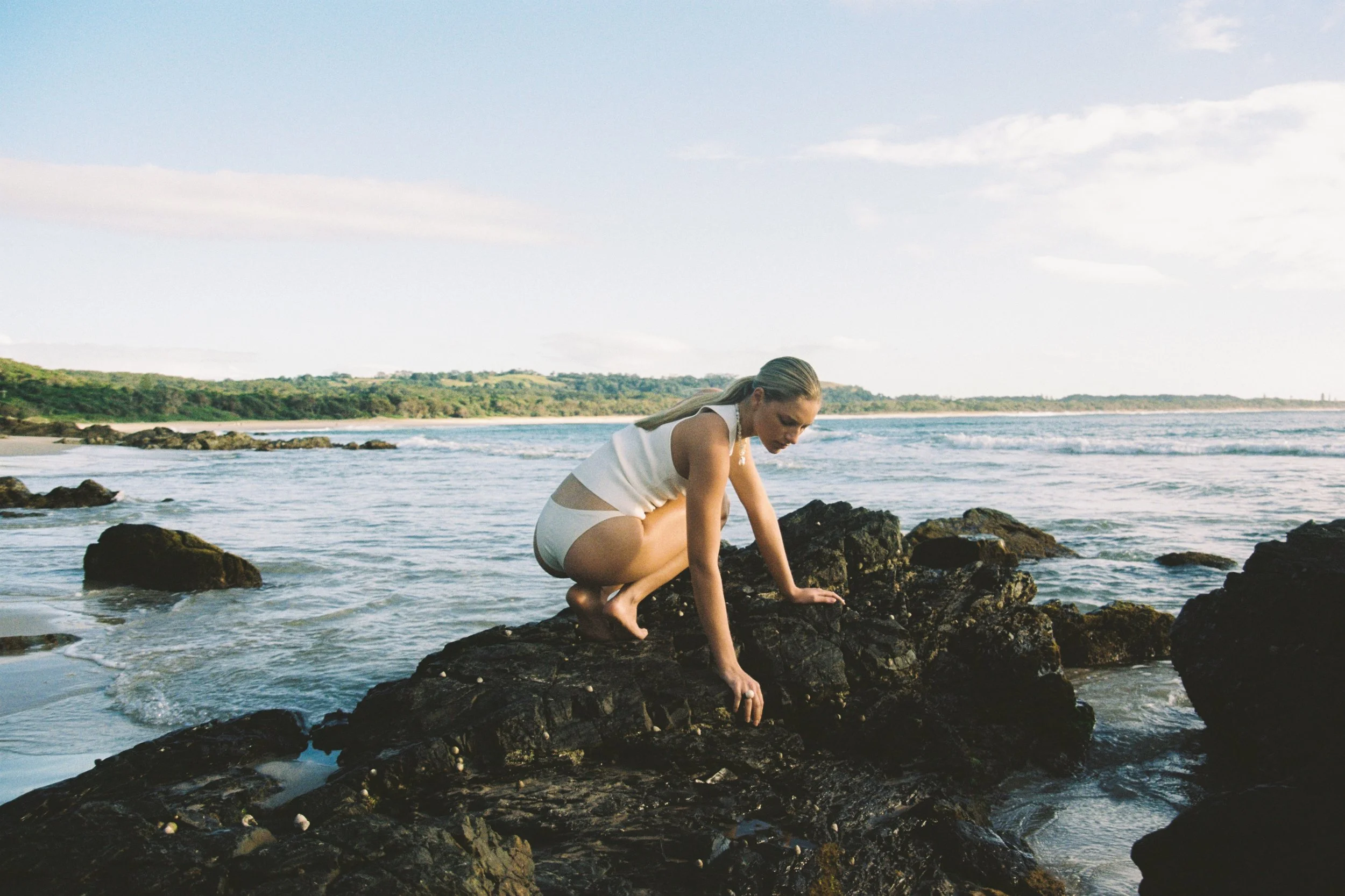 A woman in a white outfit crouches on rocks at the edge of the ocean, with waves and a distant shoreline visible in the background.