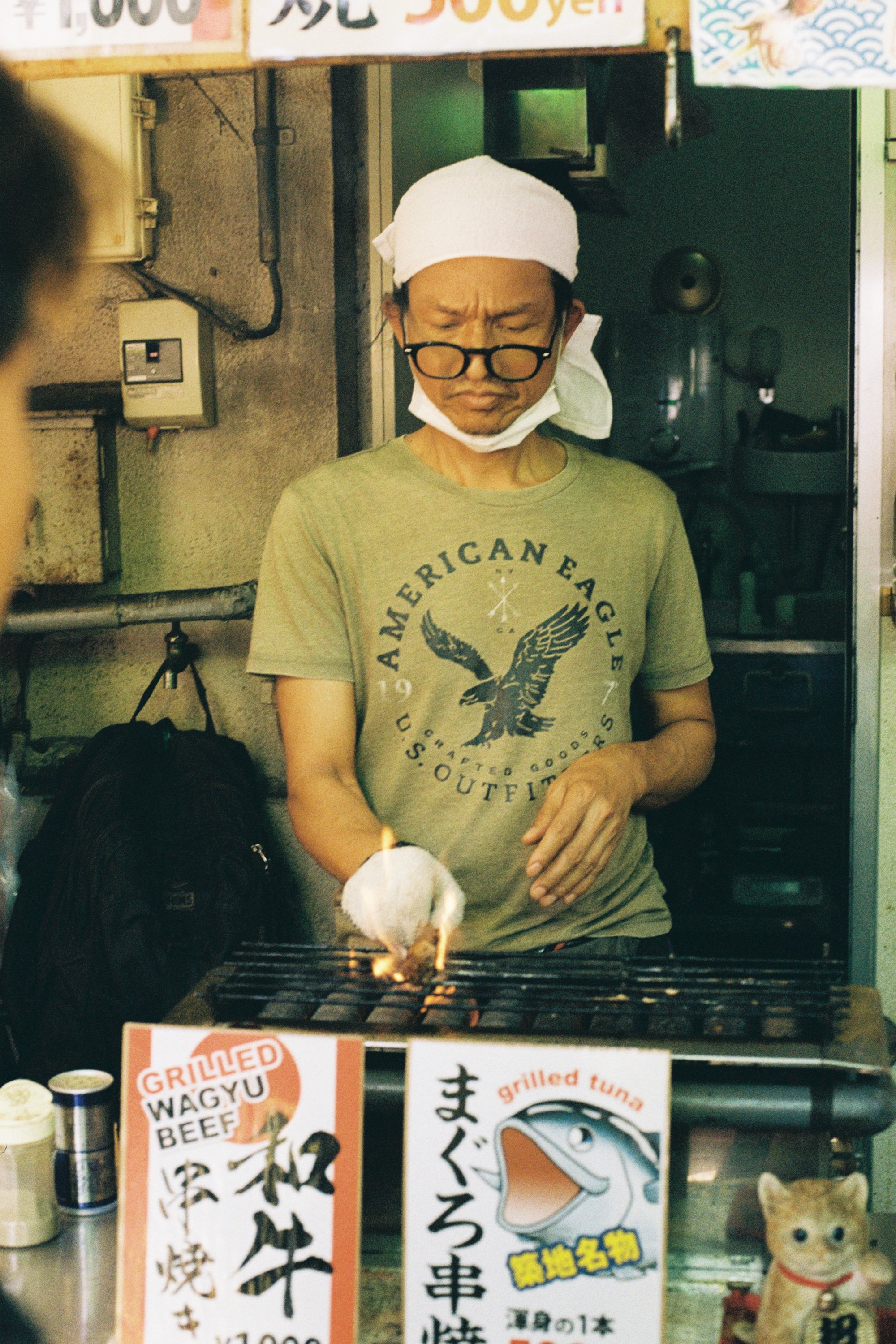 A man wearing glasses and a white headscarf is cooking over a grill, with a flame and a piece of food on the grill in front of him. There are signs with Japanese characters and some English words like 'Grilled Wagyu Beef' and 'Grilled Tuna' near the 