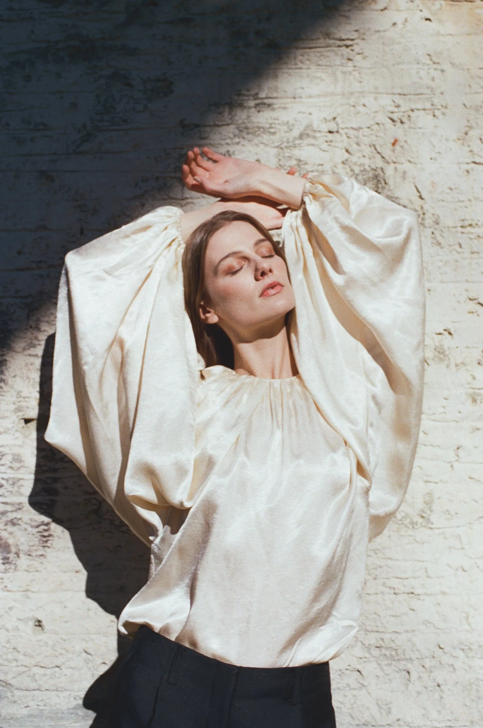 A woman with closed eyes and a relaxed expression, standing against a textured white brick wall, wearing a shiny, cream-colored blouse with voluminous sleeves and black pants, with her hands resting above her head.