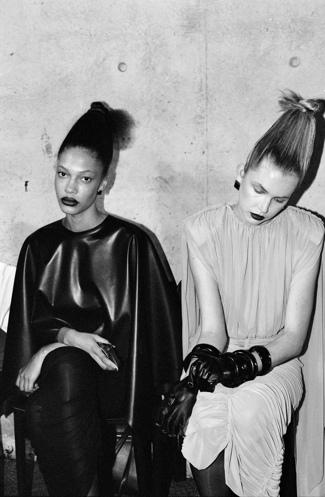 Black and white photo of two women sitting against a plain wall, both with high, styled hair and dark lipstick. The woman on the left wears a shiny black top, and the woman on the right wears a light, ruffled dress and has her head tilted down, with 