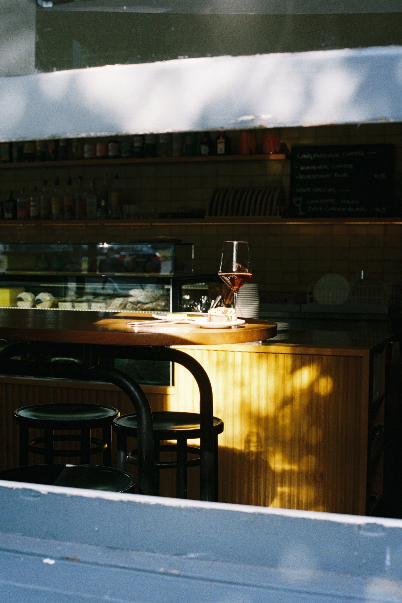 A bar with bottles of alcohol on shelves, a blackboard menu, and a glass of rosé wine on a wooden table with a napkin and utensils, seen through a window with a blueish frame.